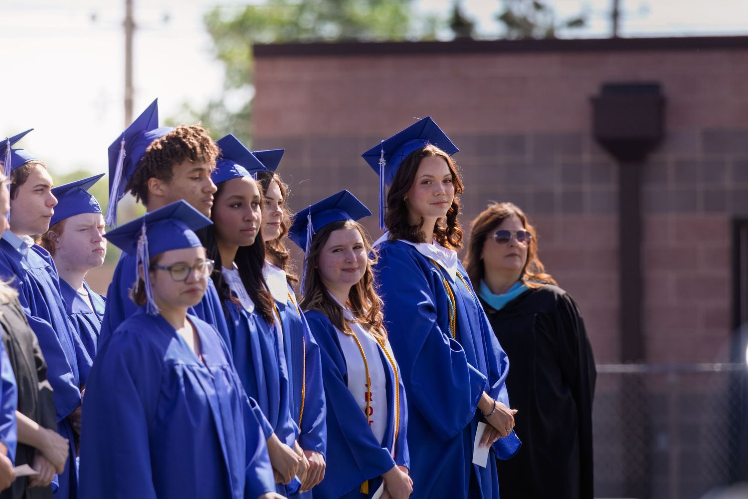 A group of graduates in bright blue caps and gowns stands together during an outdoor commencement ceremony.