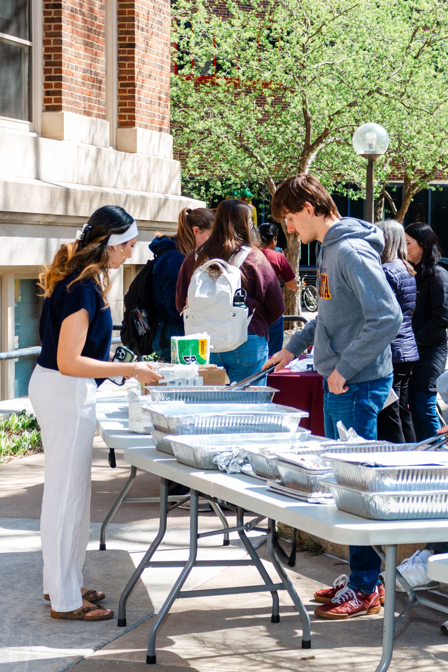 Students serving food at an outdoor campus event with foil trays on folding tables on a sunny spring day.