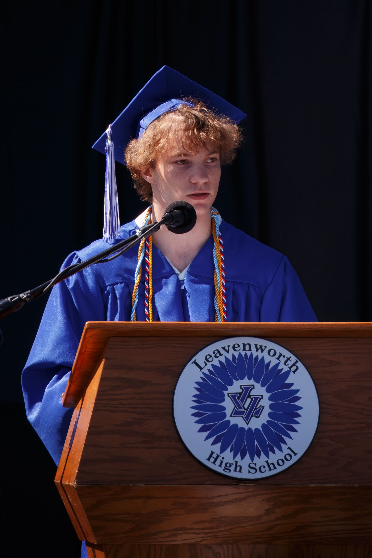 Student in blue graduation cap and gown speaks at podium with Leavenworth High School seal during ceremony.