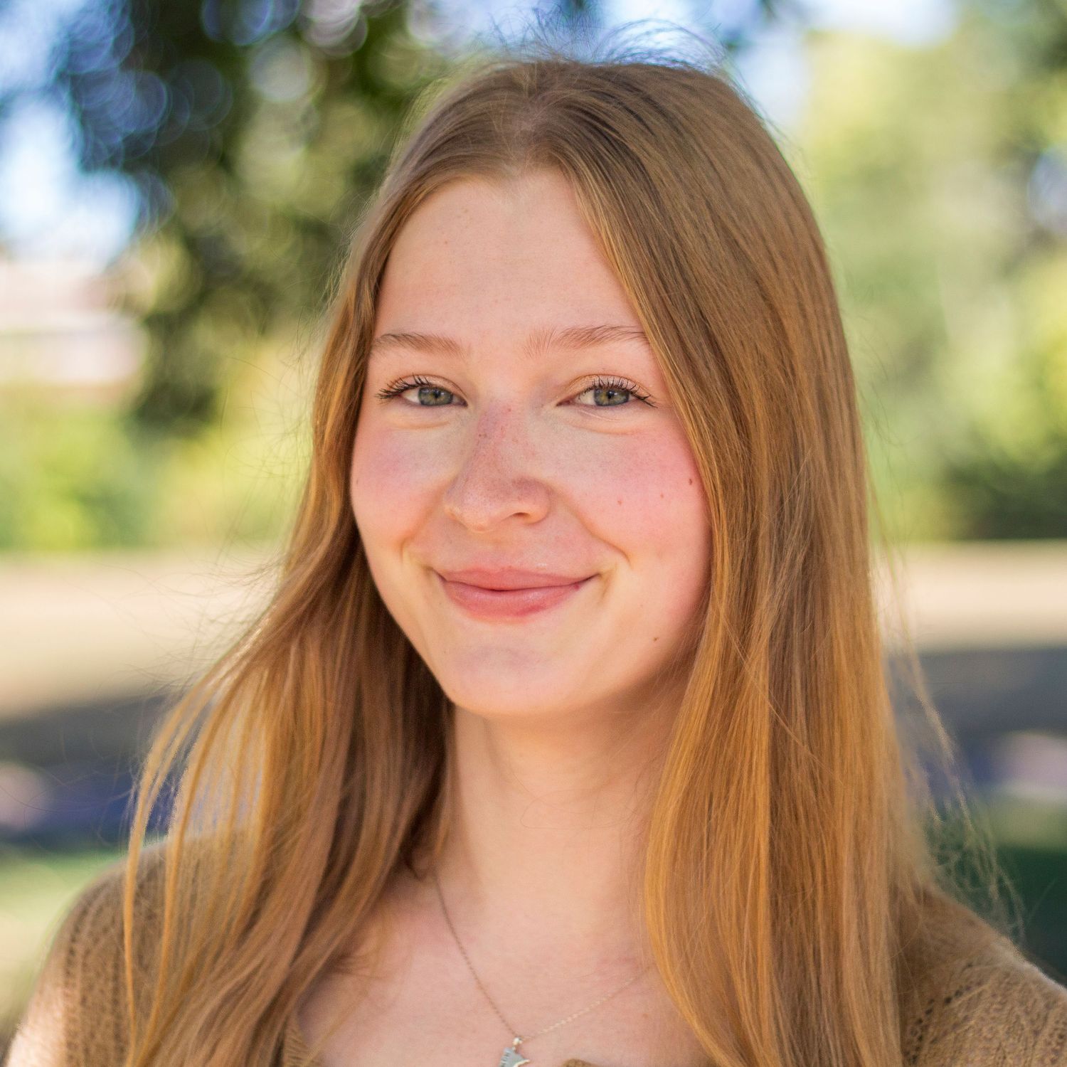 Portrait photo of someone with long straight brown hair and a warm smile outdoors with sunlit bokeh background.
