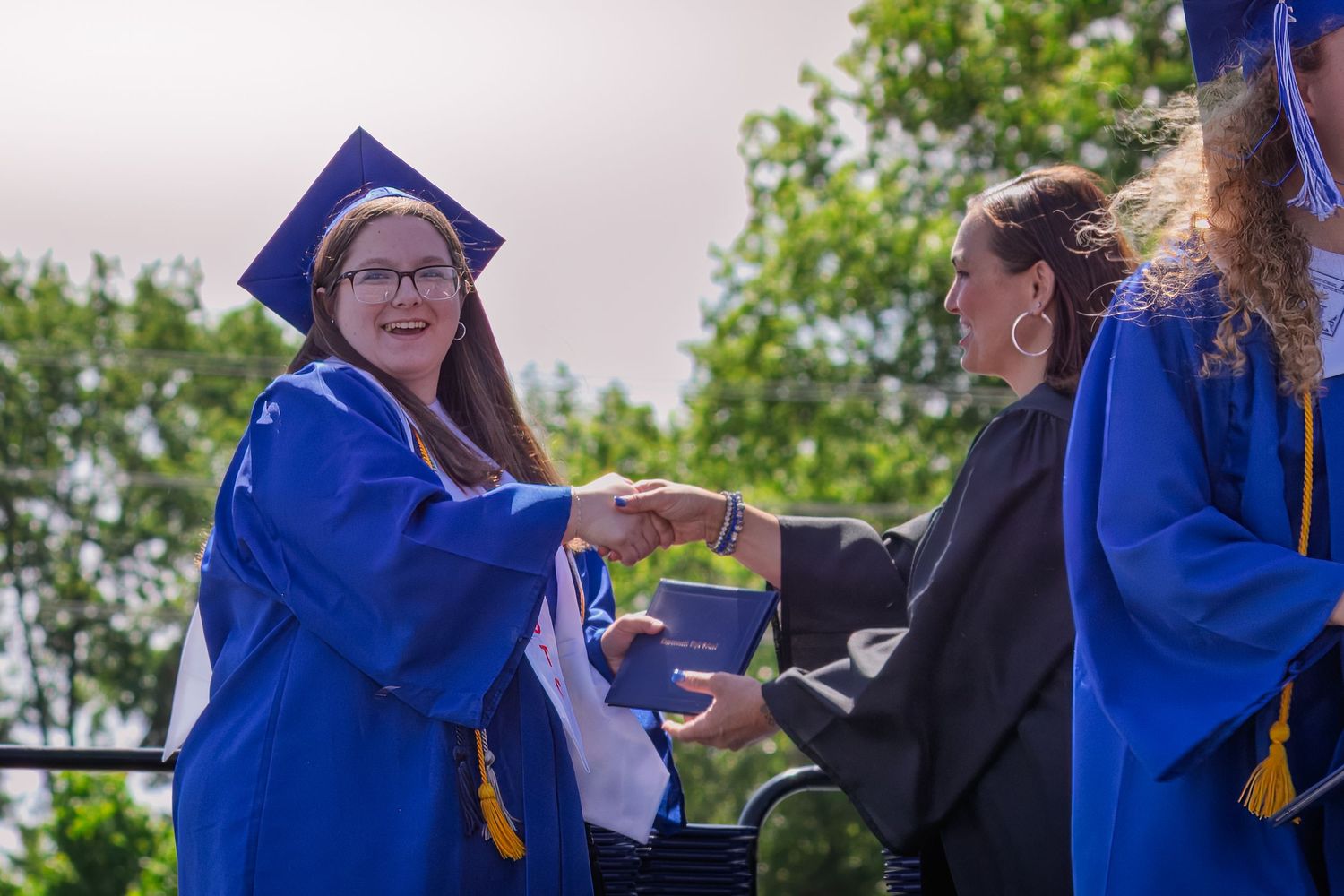 Graduate in blue cap and gown receives diploma during outdoor commencement ceremony on cloudy day.