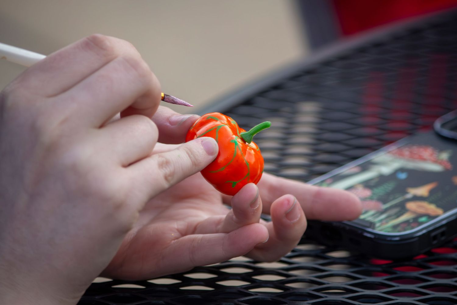 A hand holds a small red cherry tomato against a dark metal table surface with a mobile phone nearby.