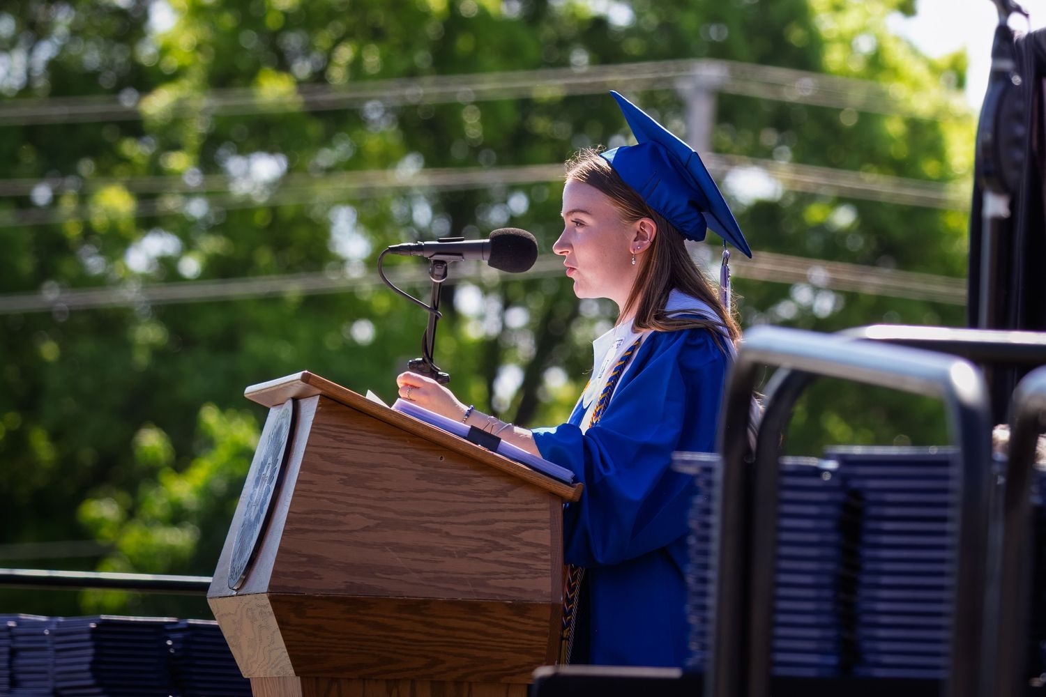 Graduate in bright blue cap and gown delivers speech at outdoor commencement ceremony from wooden podium.