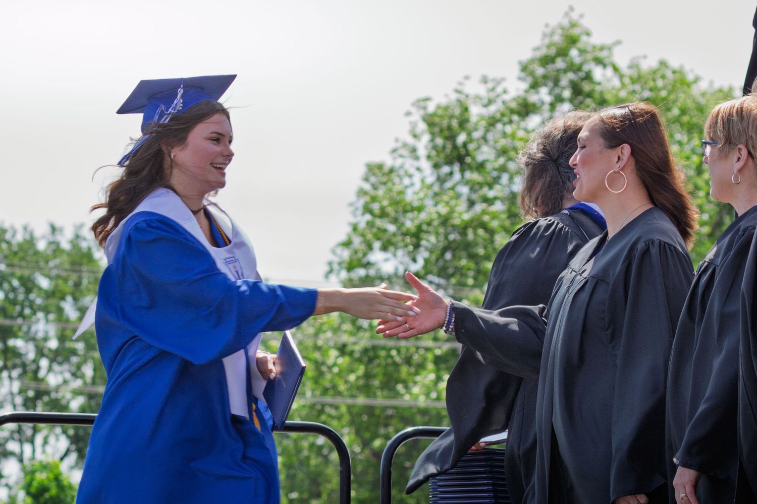 Graduate in blue cap and gown reaches to receive diploma from faculty members at outdoor ceremony.