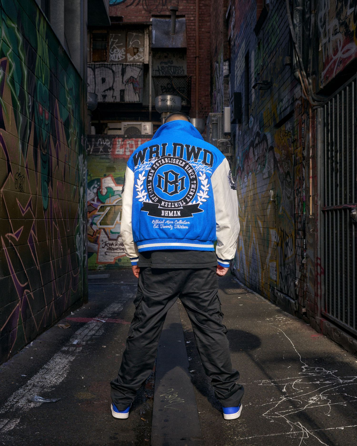 Person wearing blue and white WRLDWD varsity jacket with black pants in a graffiti-covered urban alley.