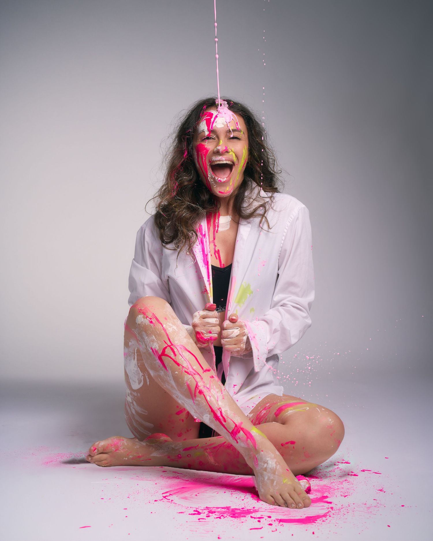 Woman covered in pink paint laughing with mouth open, sitting cross-legged in white shirt as paint pours down.