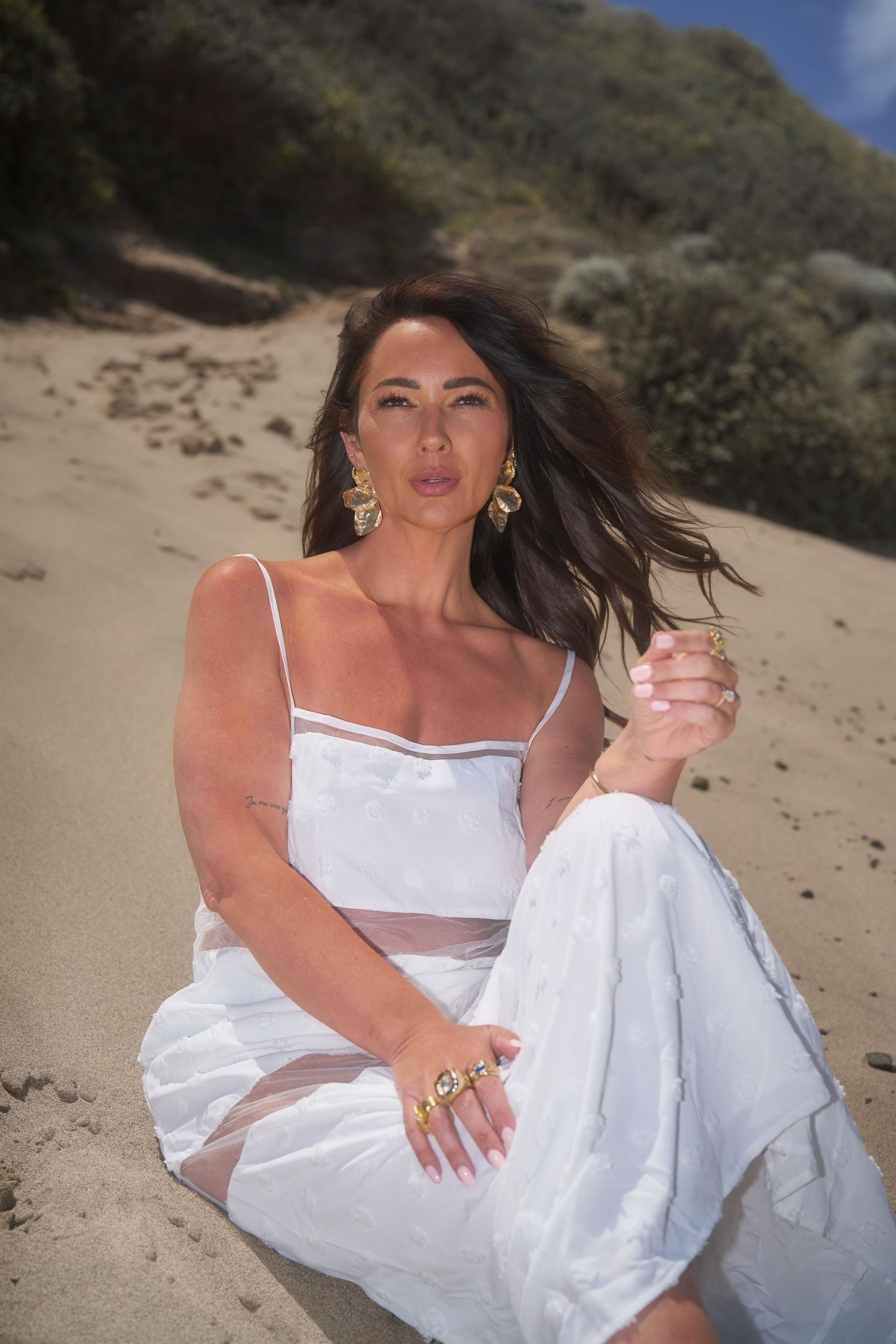 Woman in flowing white dress poses gracefully on sandy beach during outdoor photoshoot.