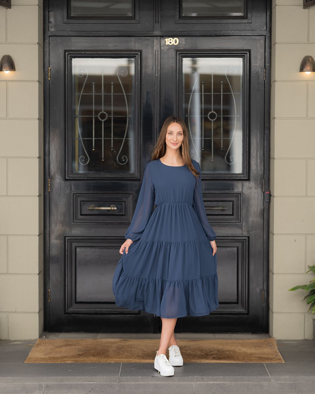 A person in a navy tiered midi dress and white sneakers stands in front of a black double door with decorative windows.