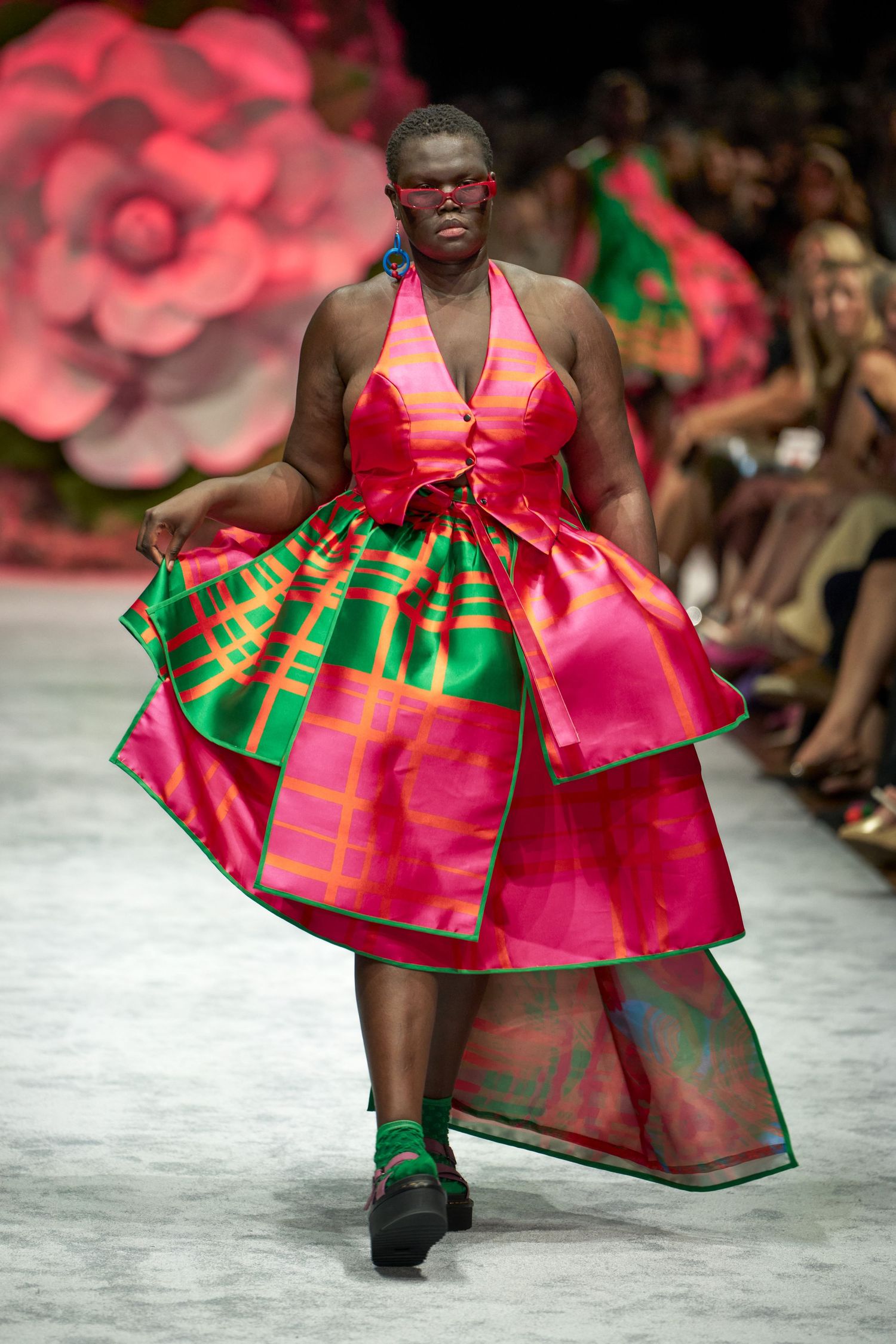 Fashion model walks runway in vibrant pink and green patterned dress with full skirt at fashion show.