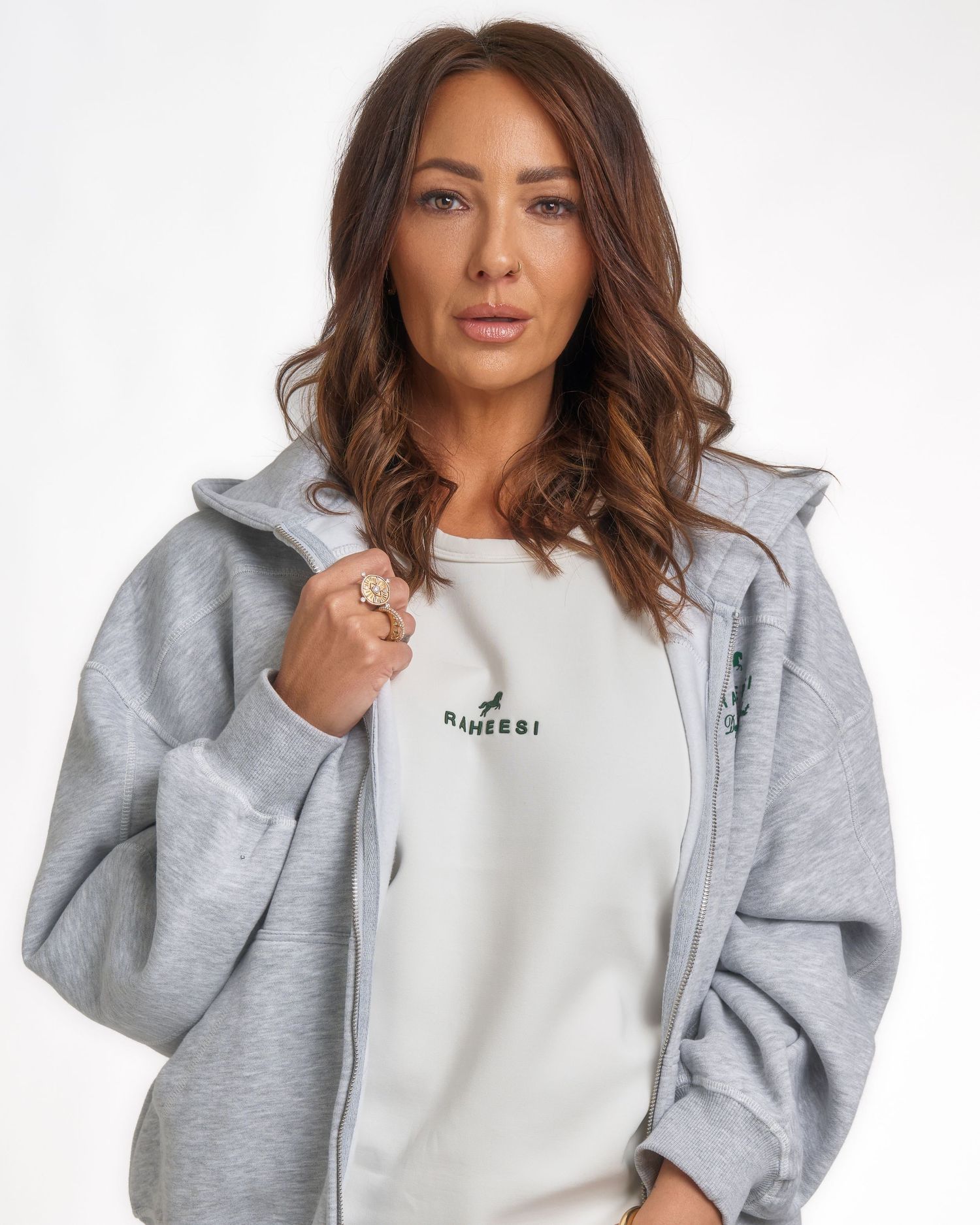 Woman in grey zip-up hoodie and white Raheesi branded sweatshirt posing against white background.