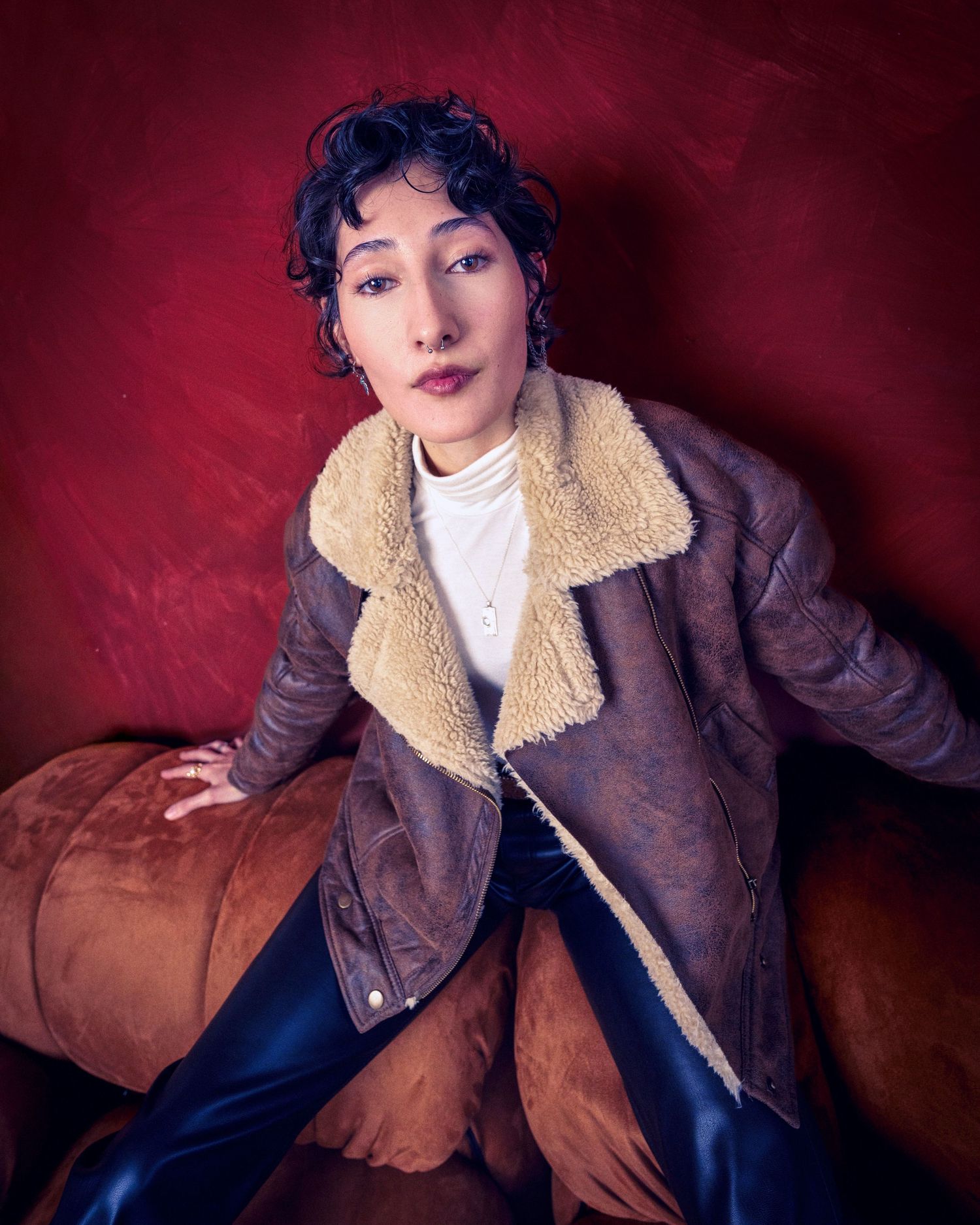 Portrait against red background wearing brown shearling jacket and white top while reclining on leather couch.