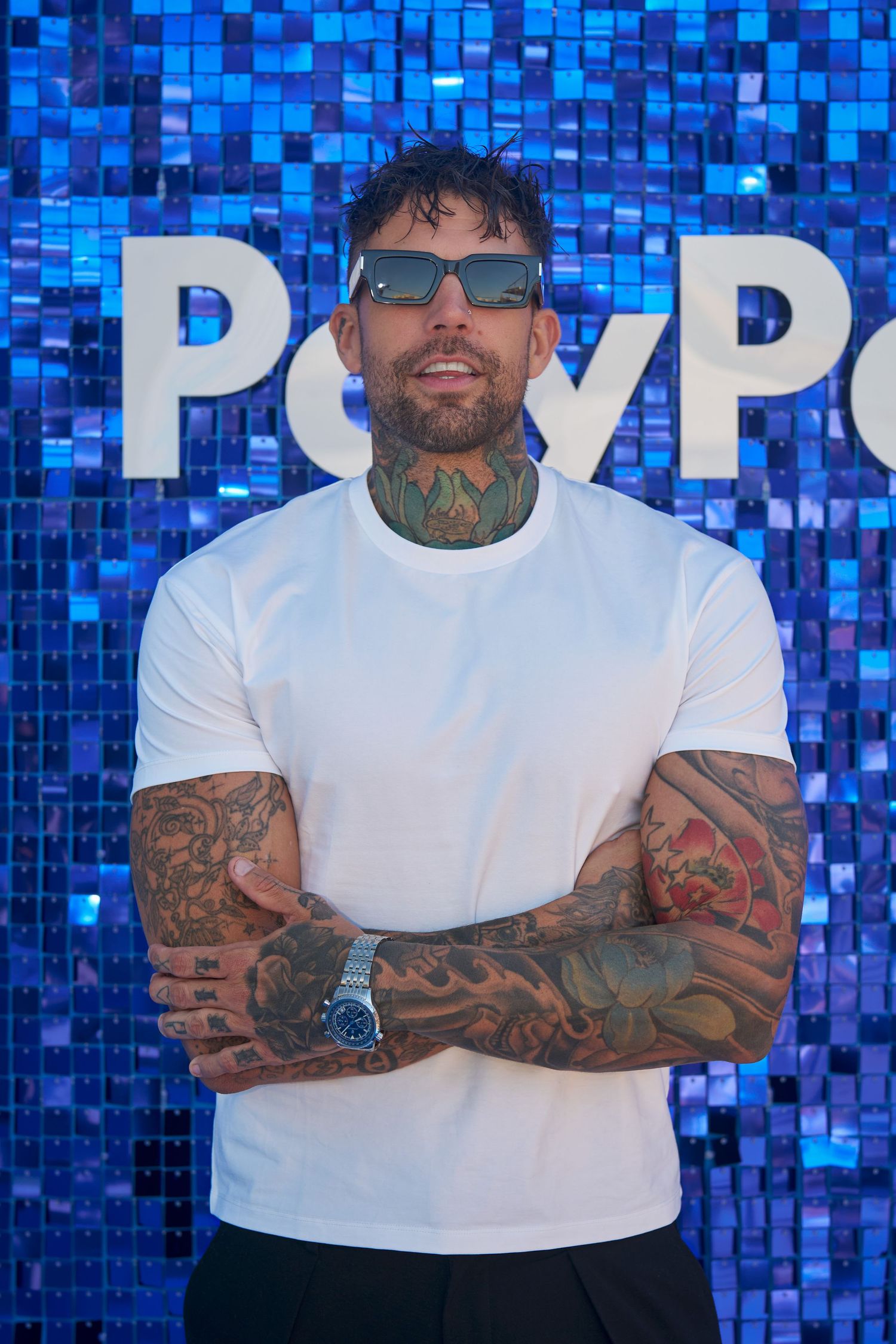 Person in white t-shirt with tattoos poses against vibrant blue mosaic tile wall.
