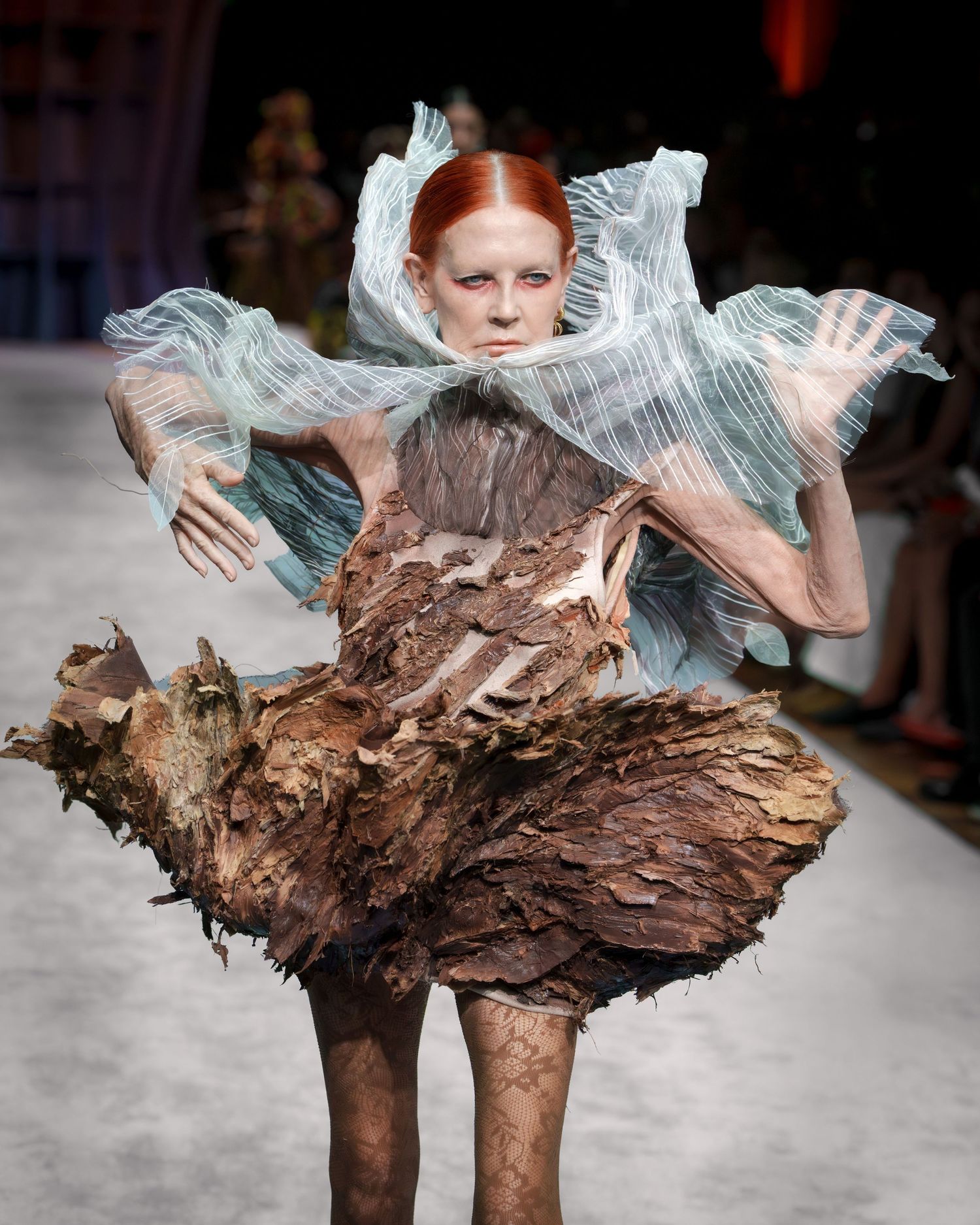 High fashion runway model wearing an avant-garde dress made of textured bark-like material with translucent ruffled details.