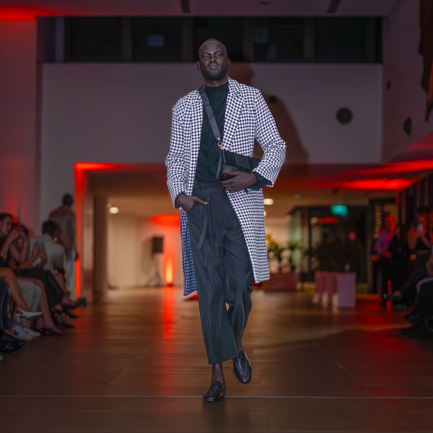 Model walks the runway wearing a checkered blazer and dark pants during a fashion show with red lighting.