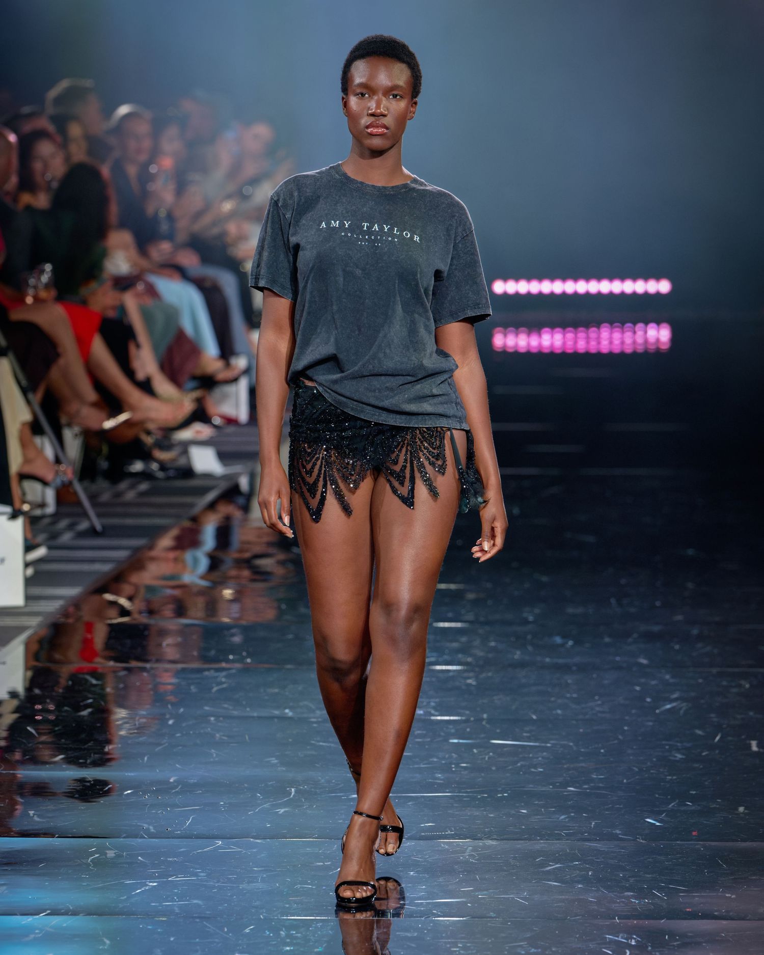 Model walks runway in gray graphic tee and black embellished mini skirt at a fashion show.