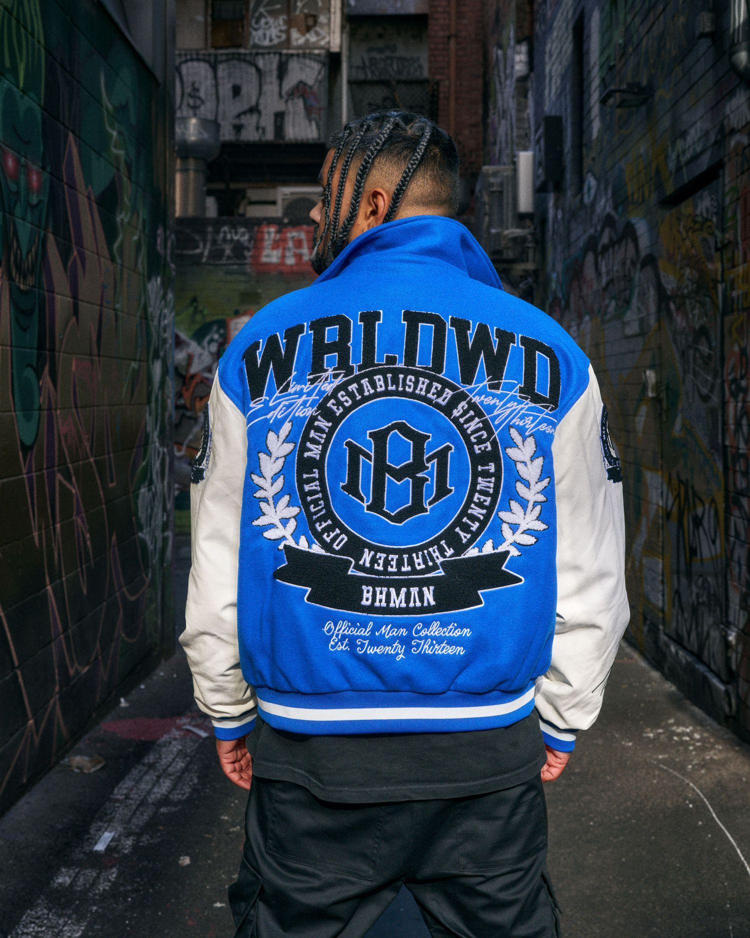 Man wearing a blue and white WRLDWD varsity jacket with embroidered logo, standing in a graffiti-lined alley.