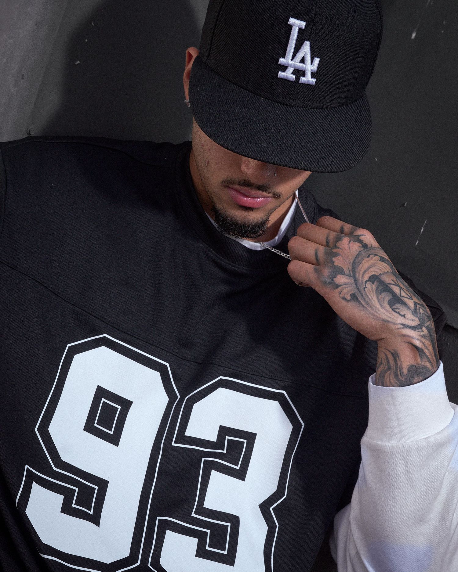 Man wearing black LA Dodgers snapback cap and number 93 jersey with tattoos on hand.