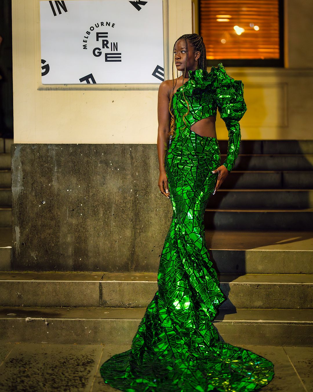 A dramatic floor-length gown made of reflective green mirror-like pieces shimmers in an industrial setting.