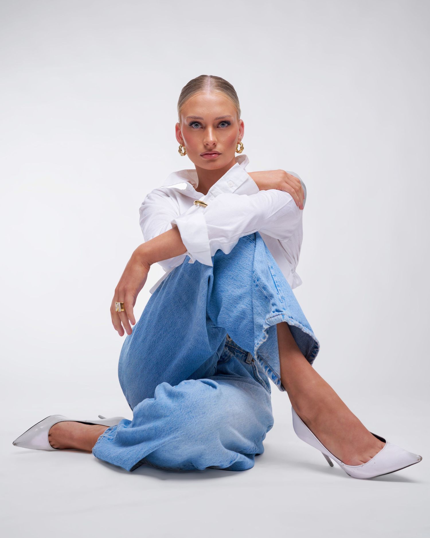 Model in casual white shirt and wide-leg jeans sits on floor wearing white heels against plain studio backdrop.