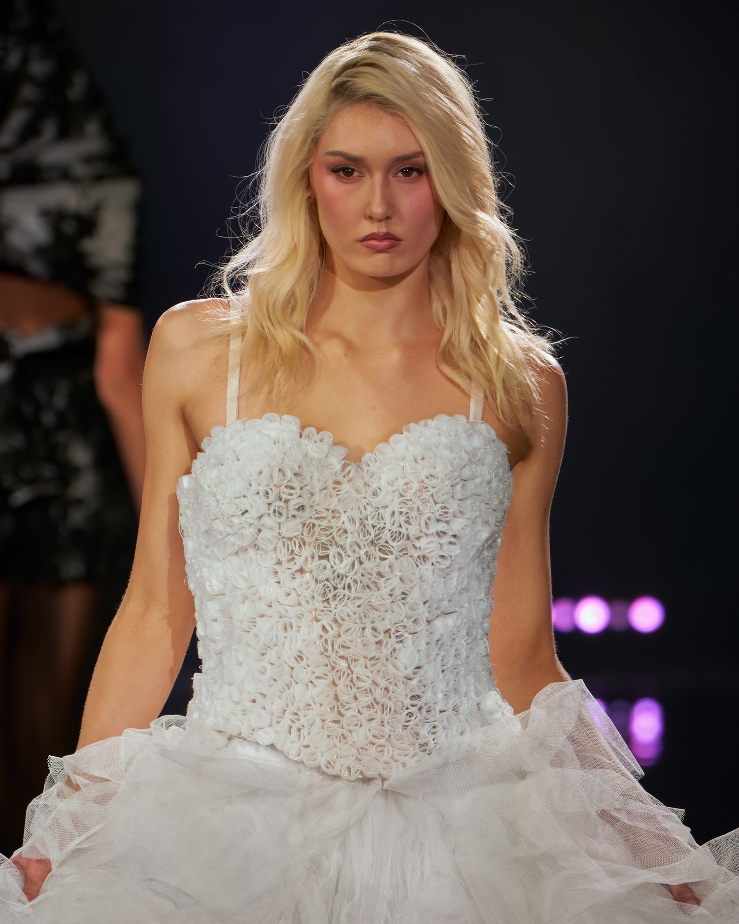 Blonde model walks runway in white lace corset bridal gown with voluminous skirt at fashion show.