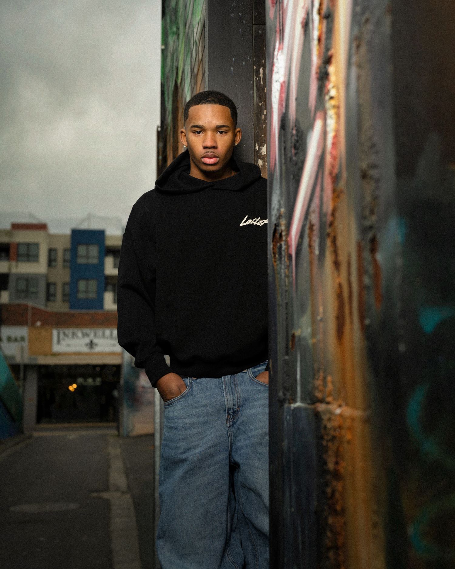 A person in a black hoodie and jeans stands against an urban wall with graffiti in moody lighting.