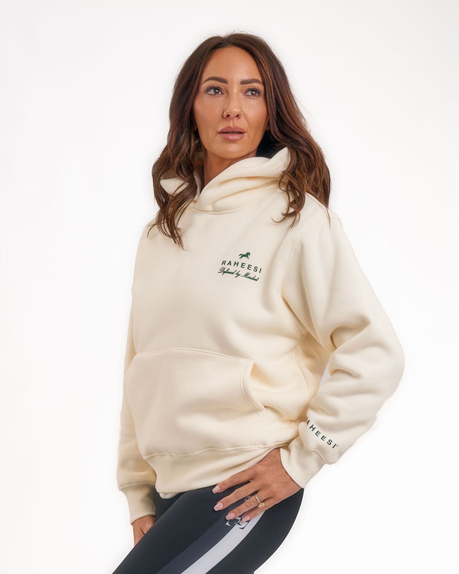 Woman wearing a cream oversized hoodie with green logo print, paired with dark leggings, posing against a white background.