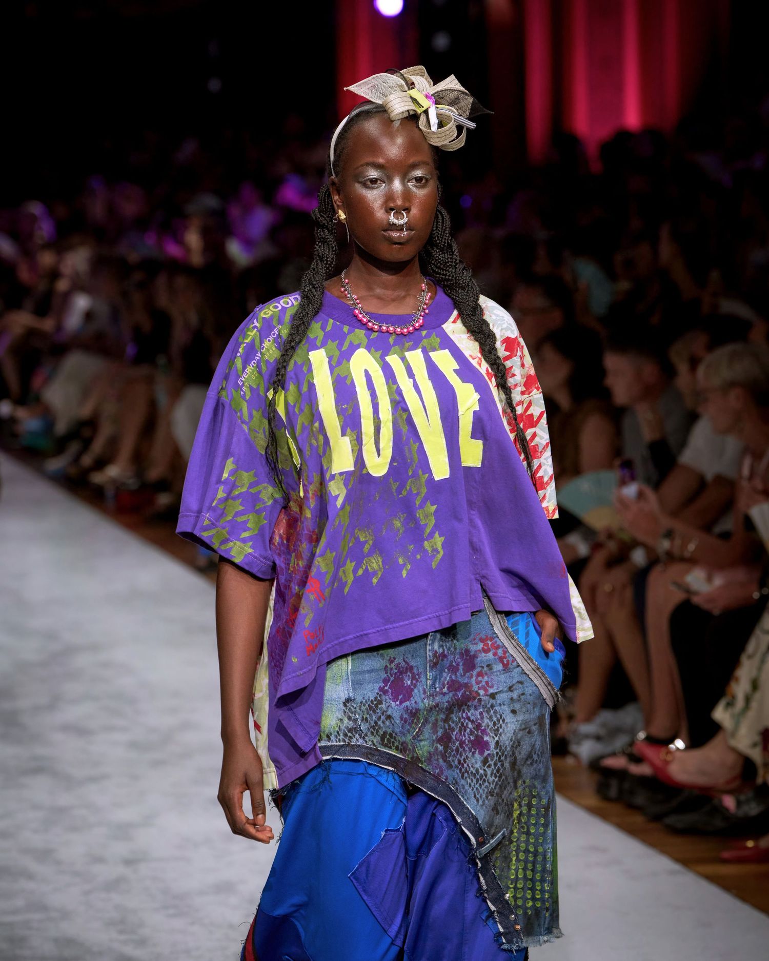 Fashion model walks runway in purple LOVE t-shirt with blue pants and floral hair accessory during fashion show.