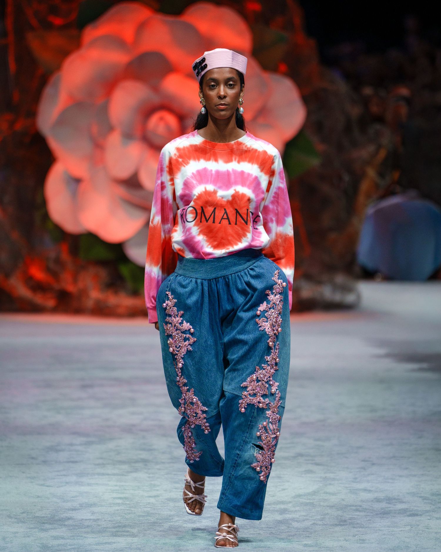 A fashion runway model wearing a red and white tie-dye sweatshirt with embroidered denim pants walks down the catwalk.