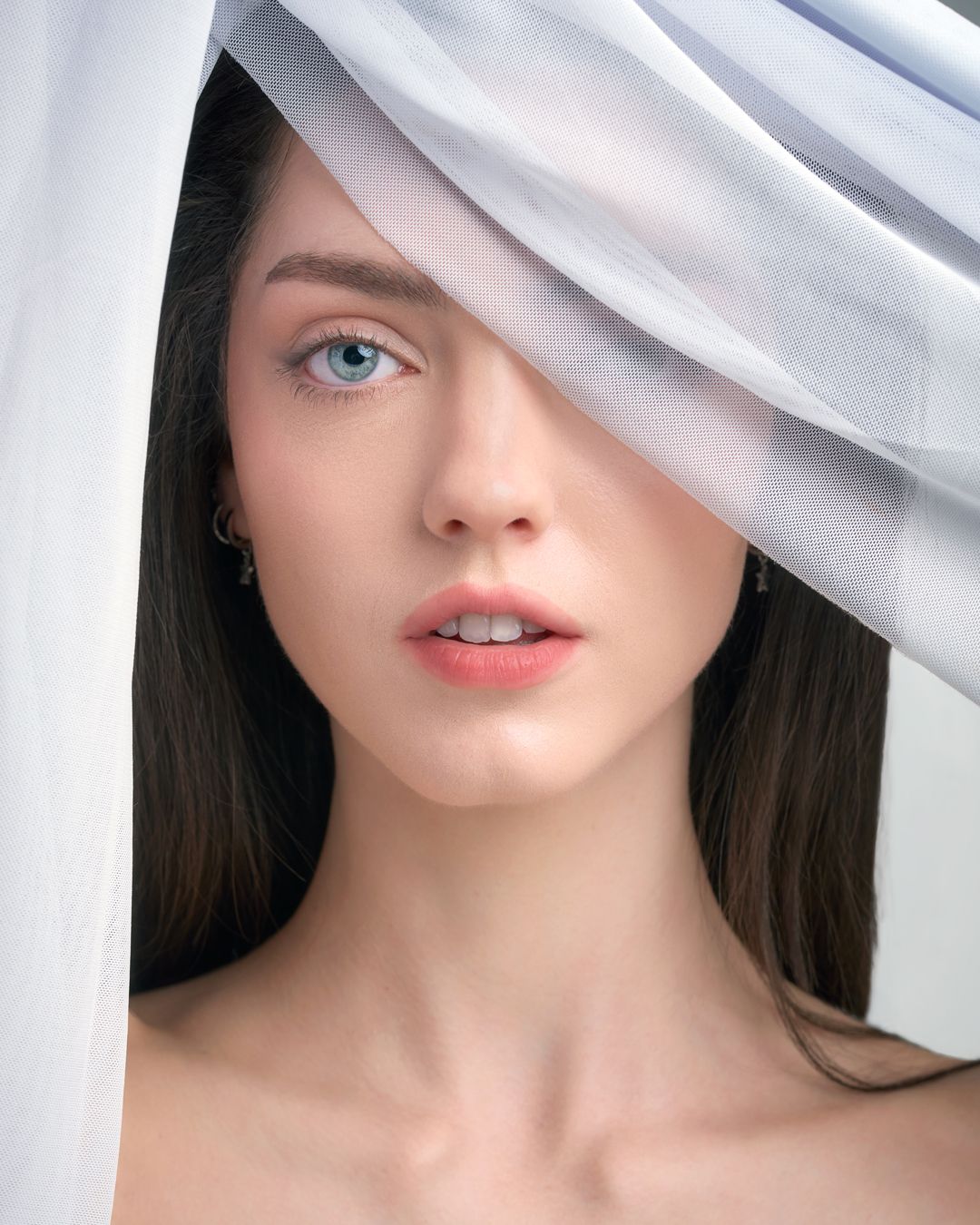 A young woman with blue eyes and dark hair peers through a flowing white sheer fabric draped over her face.