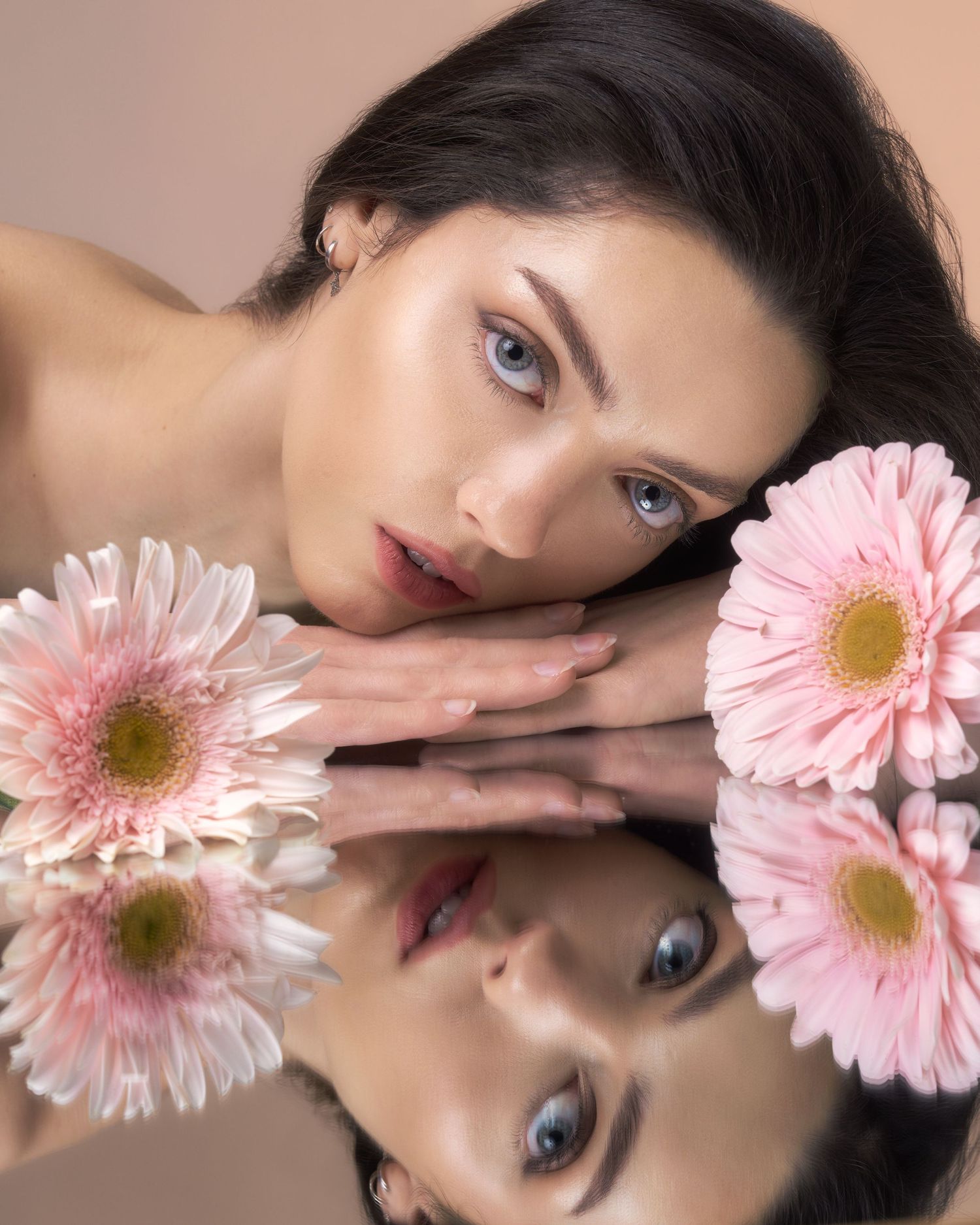 A young woman with blue eyes rests her face on a mirror surrounded by pink gerbera daisies in a beauty portrait.
