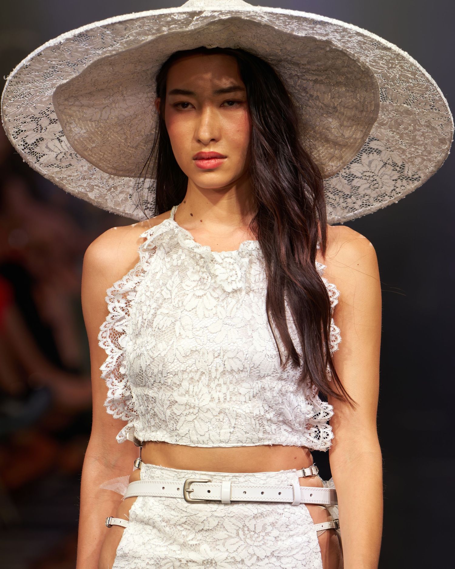 Model wearing white lace crop top, wide-brim lace hat, and white belt on a fashion runway.