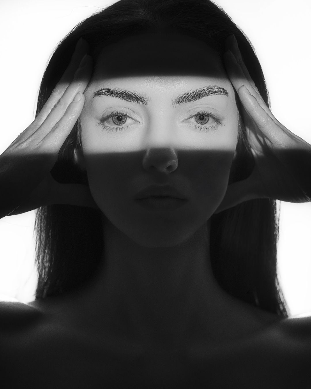 Black and white portrait of a young woman with hands on temples, dramatic lighting highlighting her striking features.