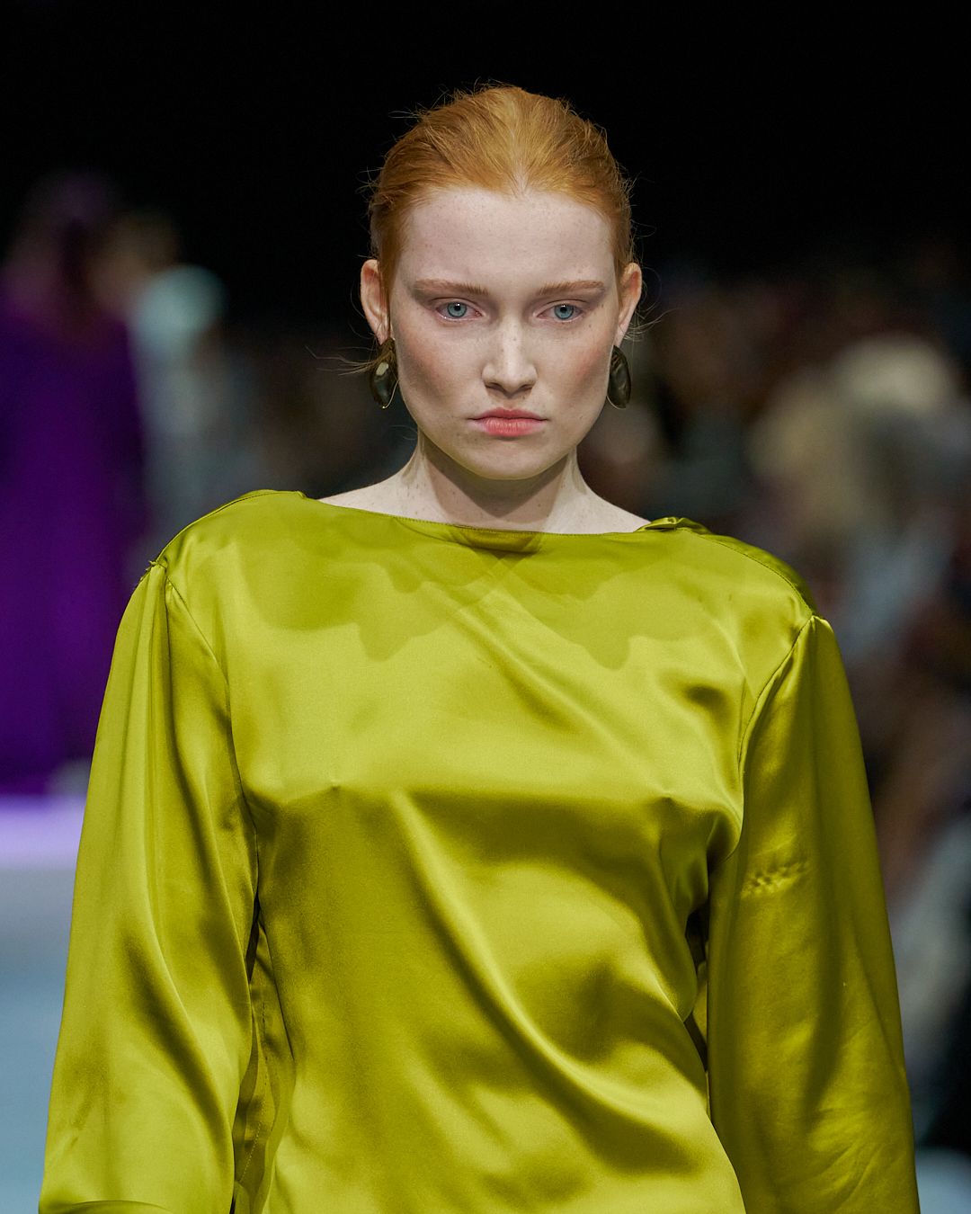 Model wearing chartreuse silk long-sleeve top on fashion runway with dark background.