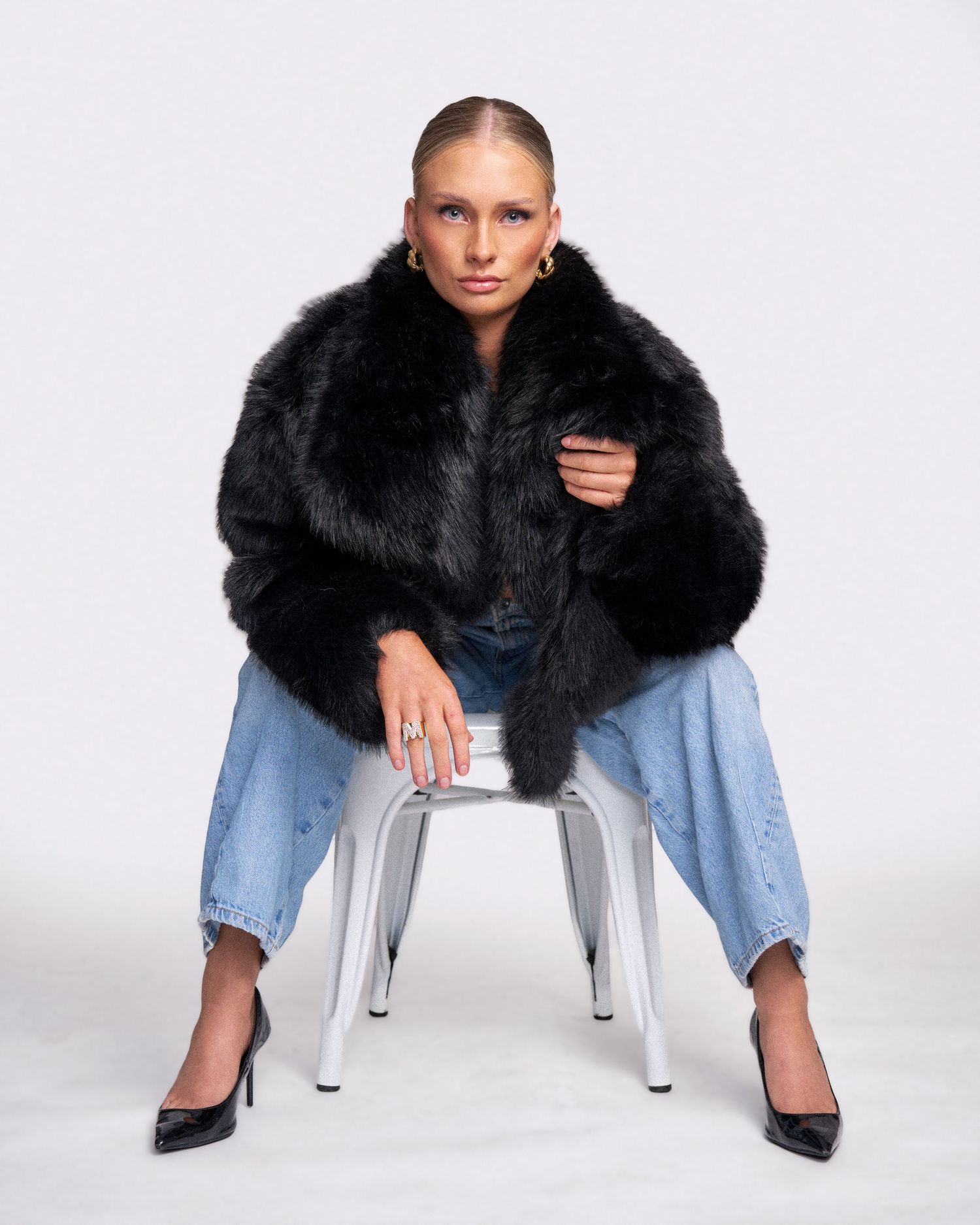 Fashion model in black fur coat and jeans sits on white stool against studio backdrop wearing black heels.