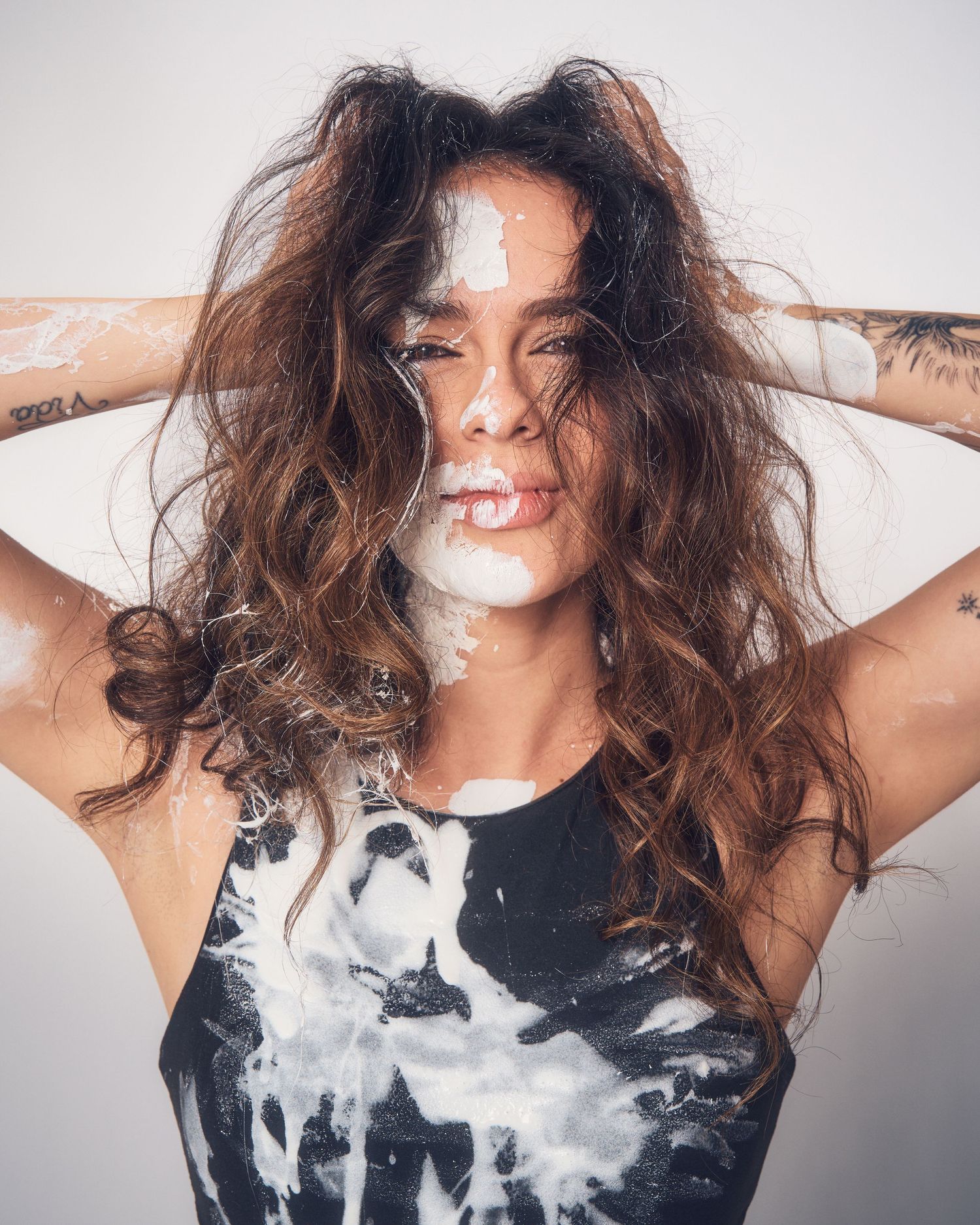 Woman with curly brown hair and tattoos poses with white paint splattered on her face and black tie-dye top.