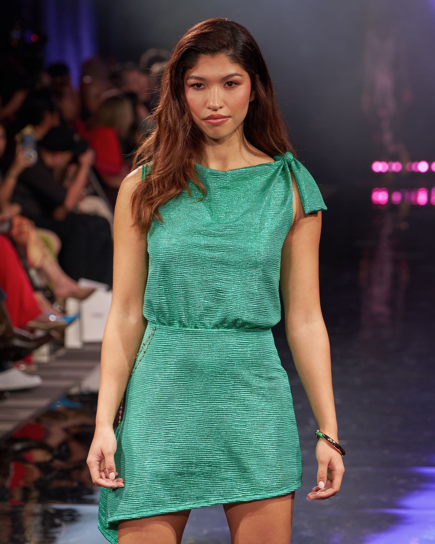 A model walks the runway in a green textured mini dress with bow shoulder detail at a fashion show.