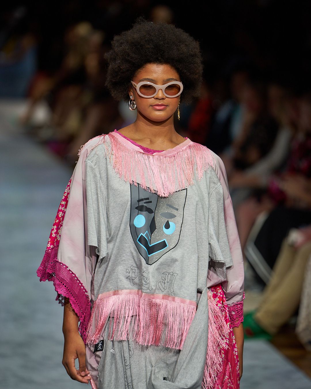 Fashion model walks runway in oversized grey t-shirt with pink fringe details and sunglasses during fashion show.