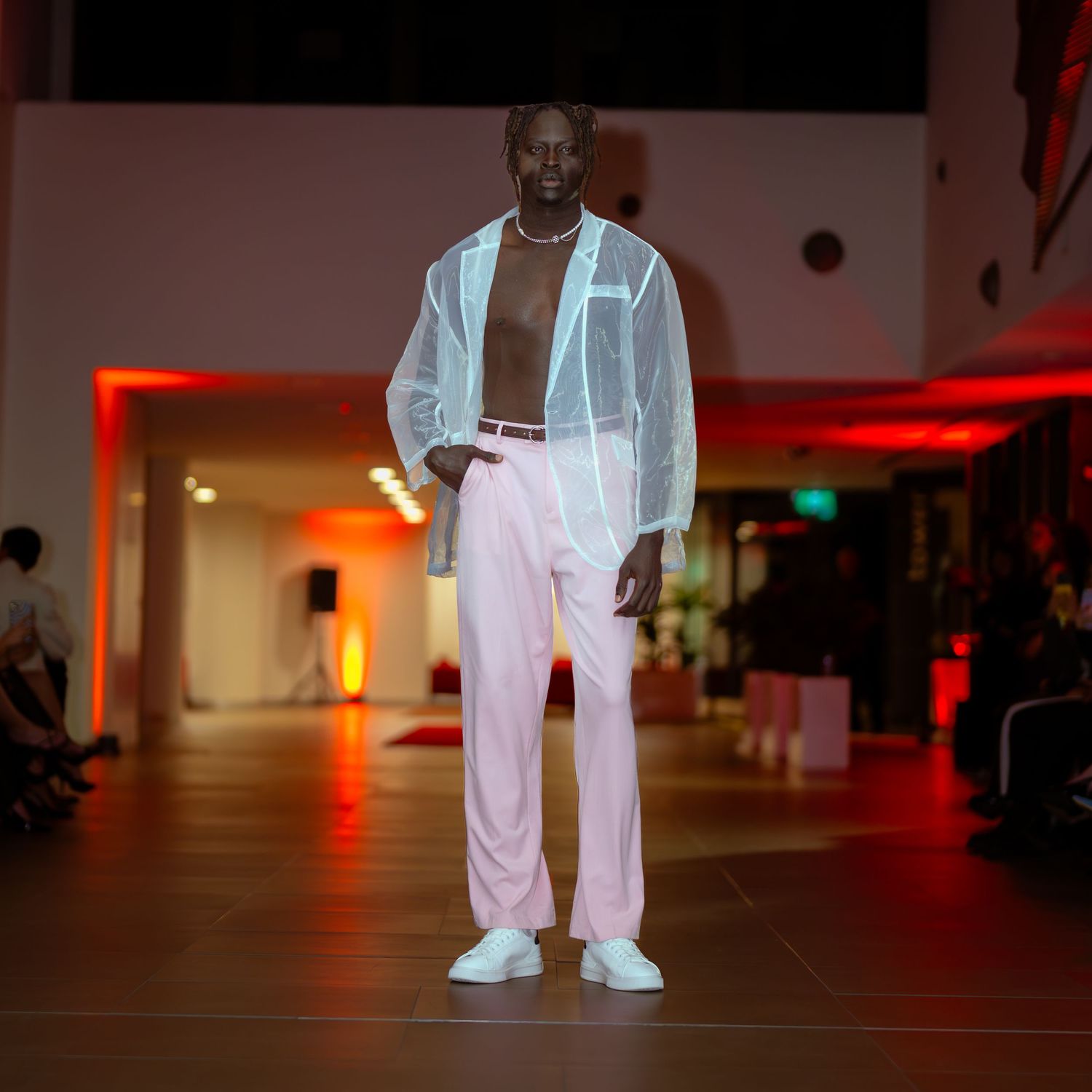 Fashion model walks runway in white oversized suit and sneakers at dimly lit fashion show.