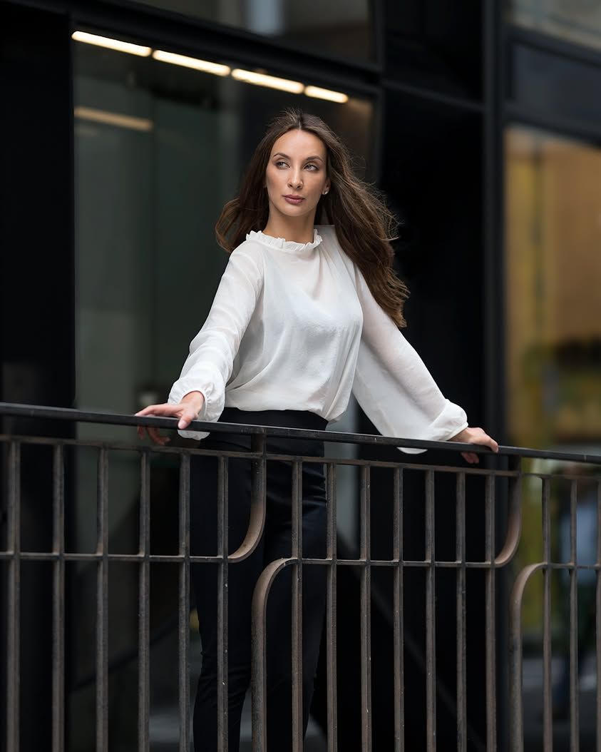 Woman in white sweater leans on metal railing while looking to the side with flowing hair in urban setting.