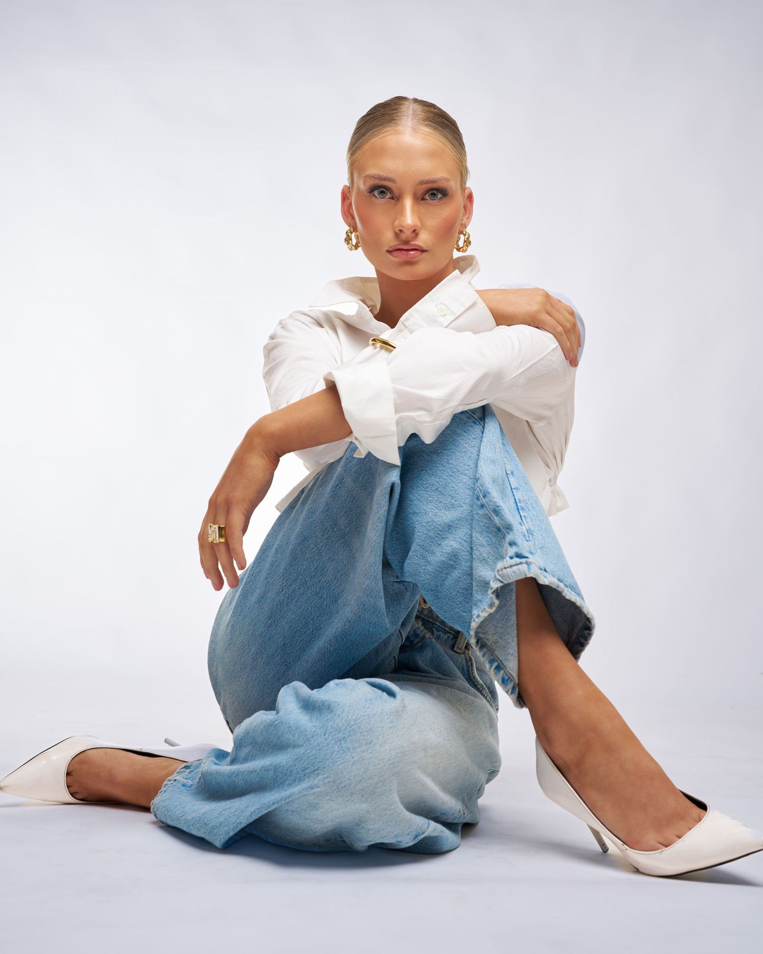 Woman in white shirt and blue jeans sitting on floor wearing white heels, posing confidently against light background.