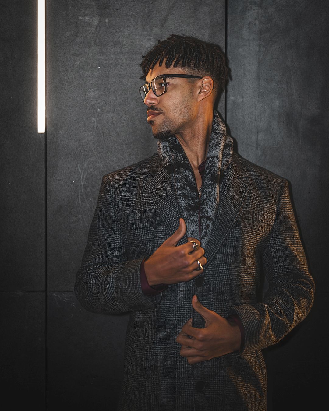 Against a dark textured wall, someone wearing glasses and a dark jacket stands in dramatic lighting in a series of poses.