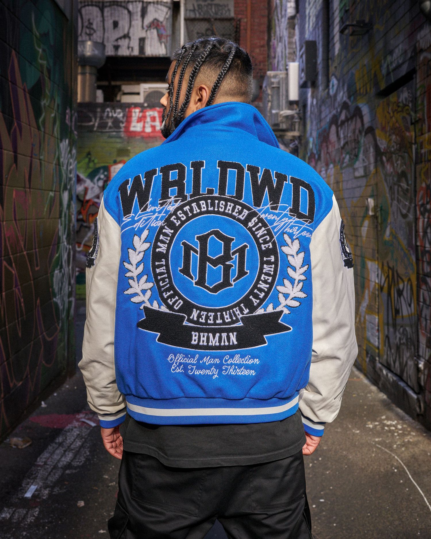 Man wearing blue and white WRLDWD varsity jacket with BHMAN logo in urban alleyway.
