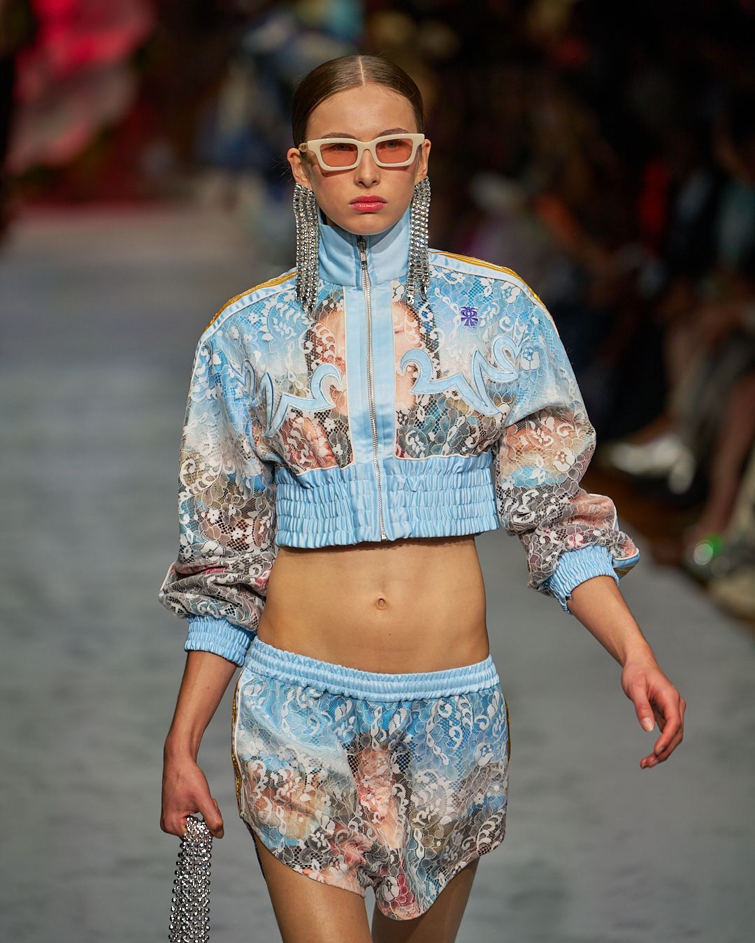 Runway model wearing a light blue and pink patterned cropped tracksuit with matching shorts and retro sunglasses.