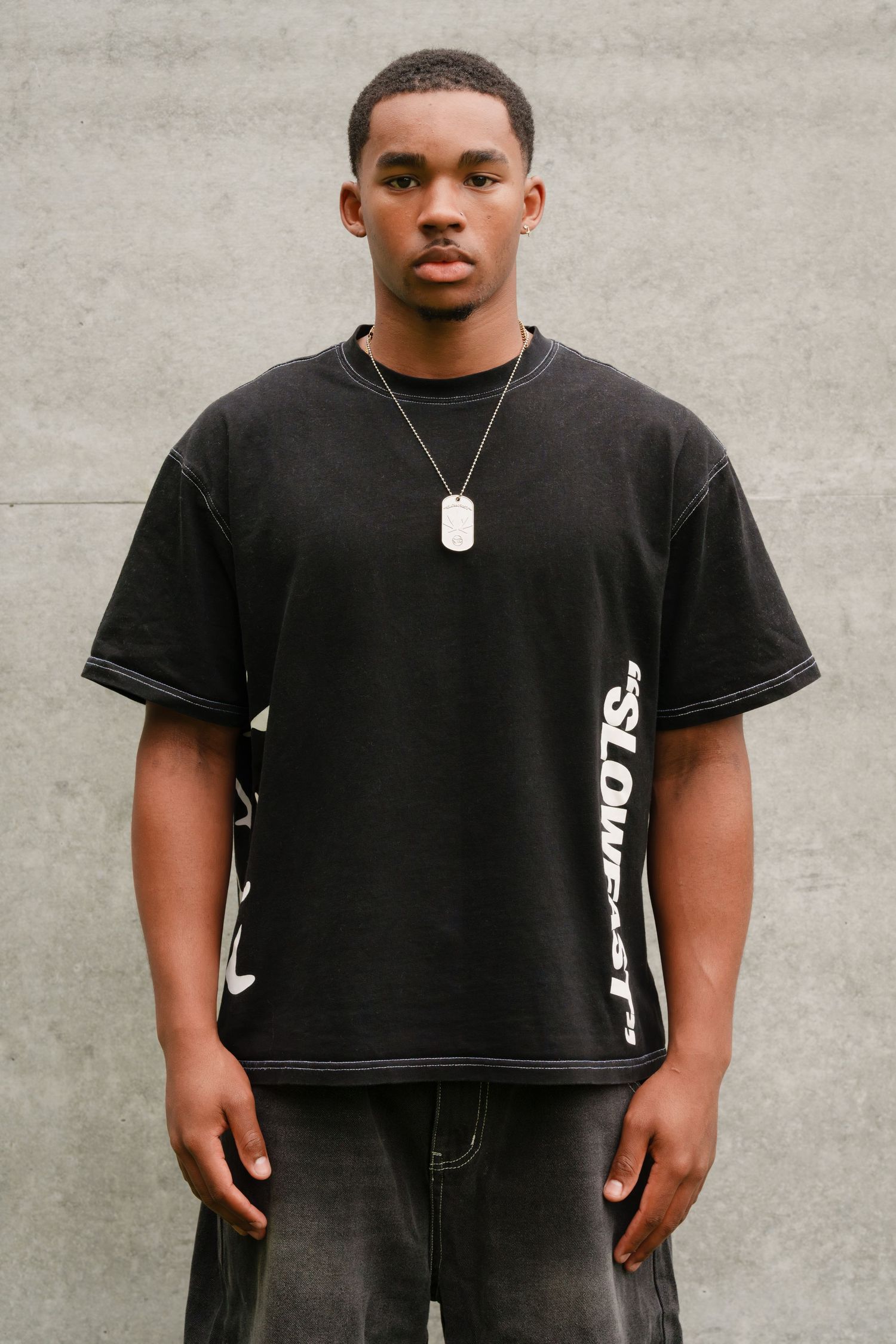Person wearing black t-shirt and shorts stands against gray wall in fashion lookbook style photo series.