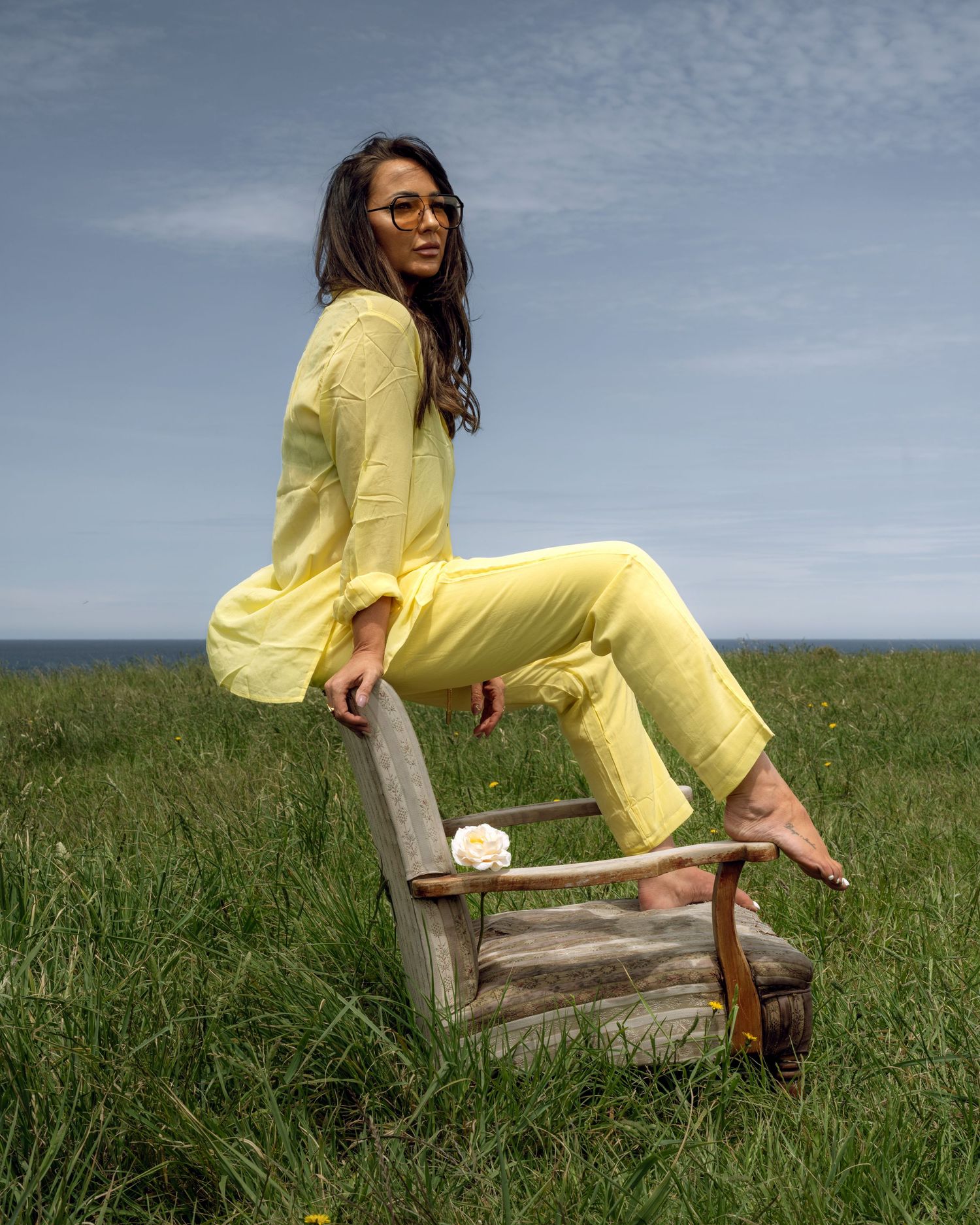 A person in a bright yellow outfit lounges on a wooden bench in a grassy field against a cloudy sky.