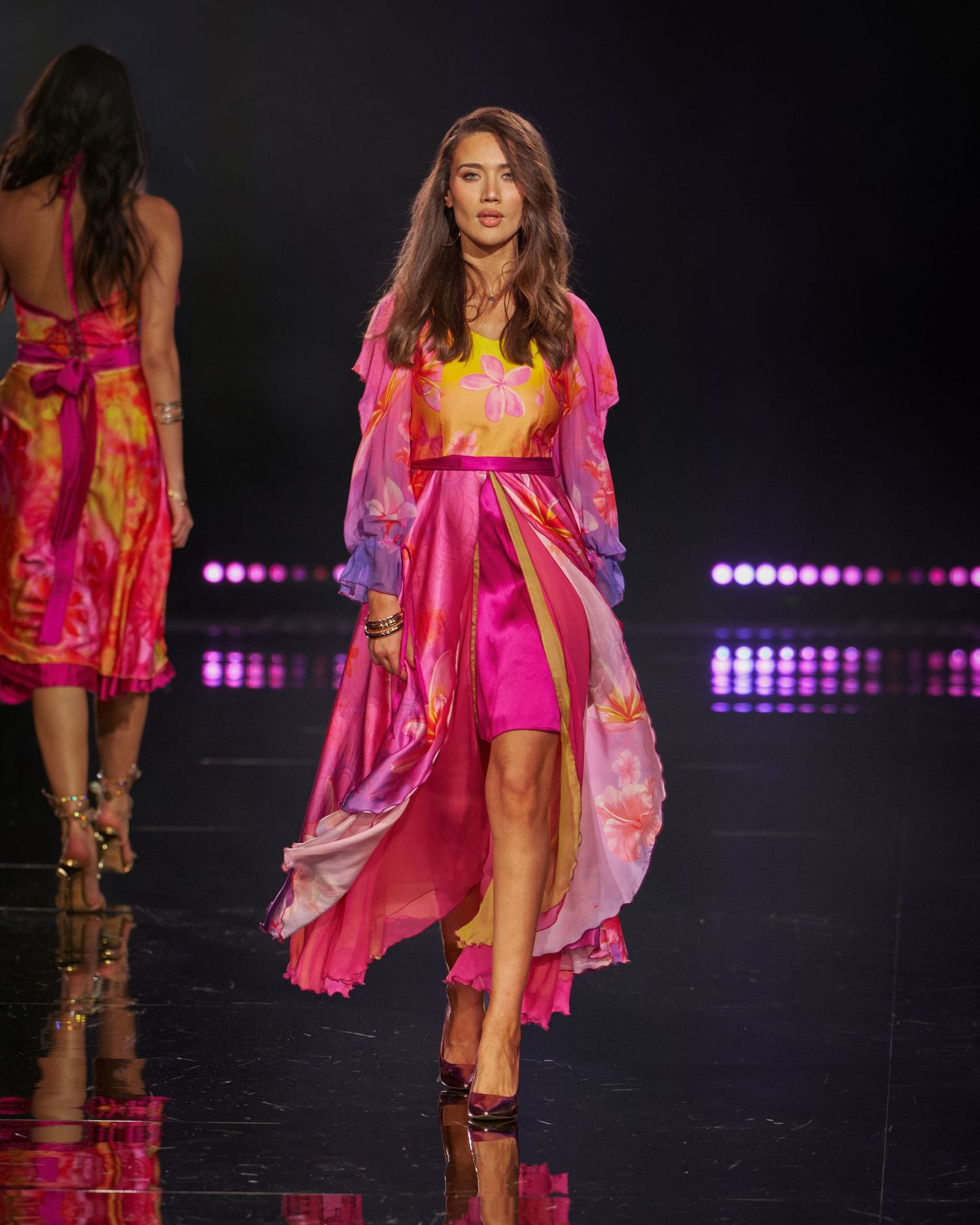 Model walks runway in vibrant pink floral maxi dress with sheer overlay at fashion show.