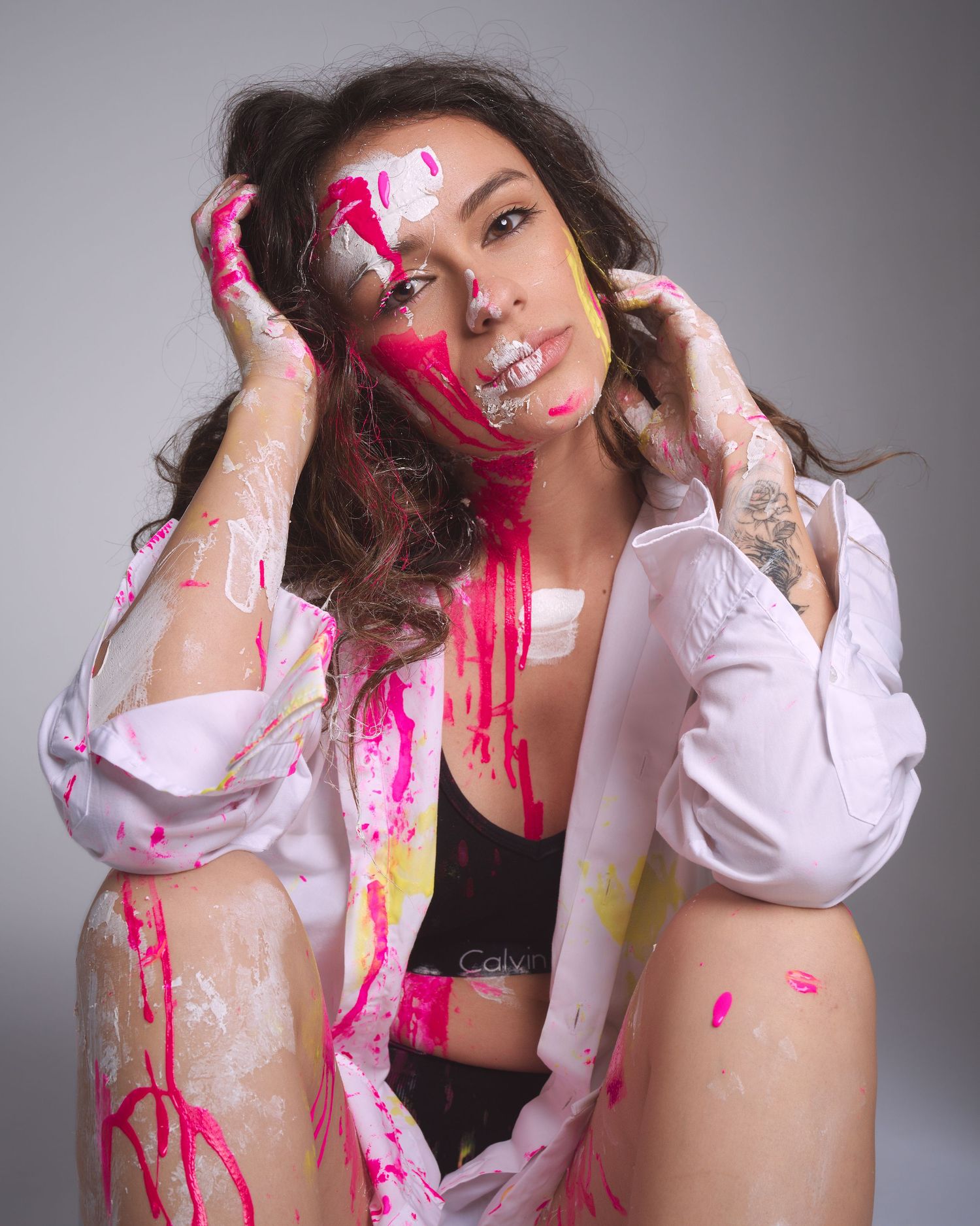 Woman covered in vibrant pink and yellow paint, wearing a white shirt, posing artistically in a studio setting.