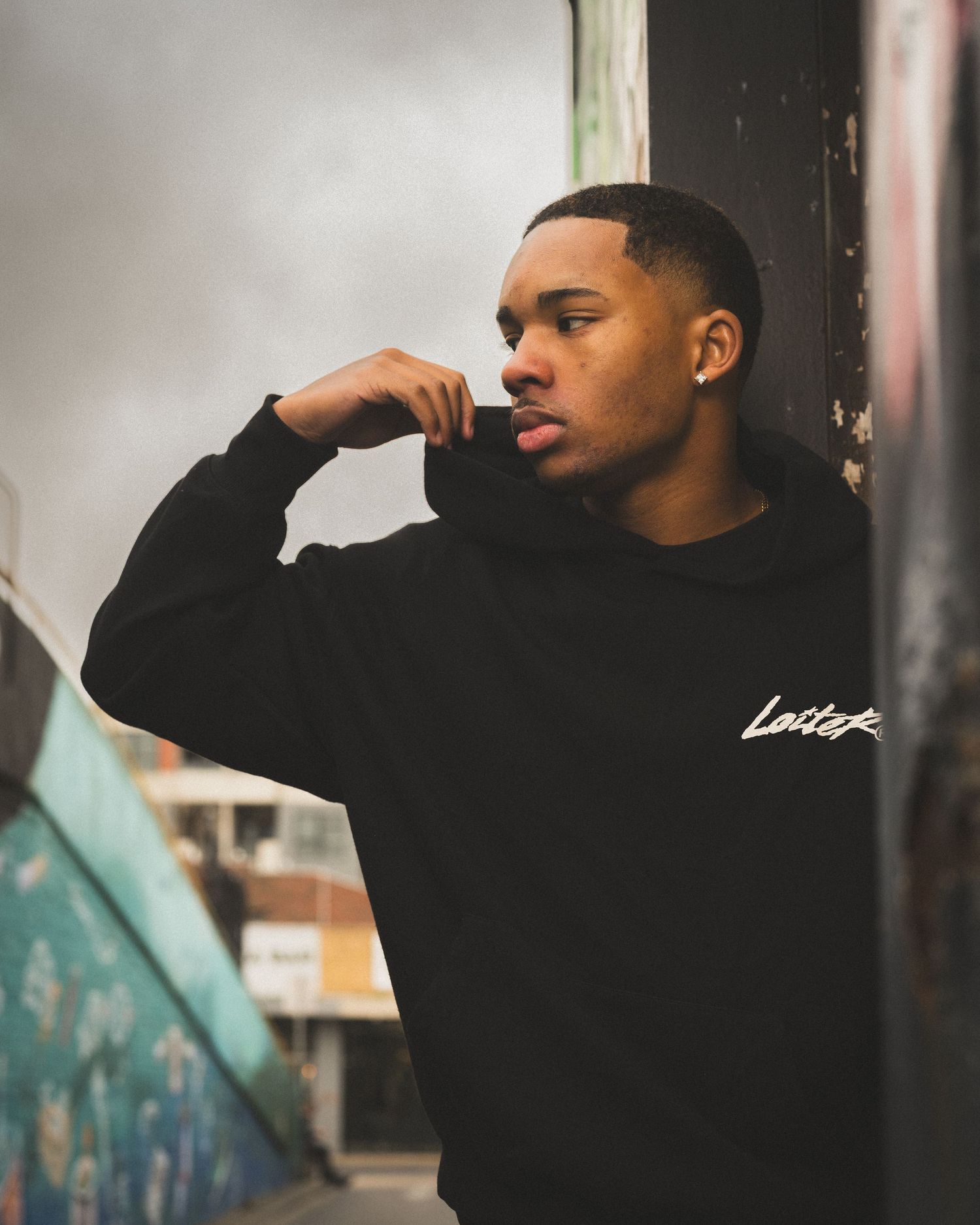 Person in black sweater with 'Laiza' text poses thoughtfully against graffiti wall in artistic portrait shot.