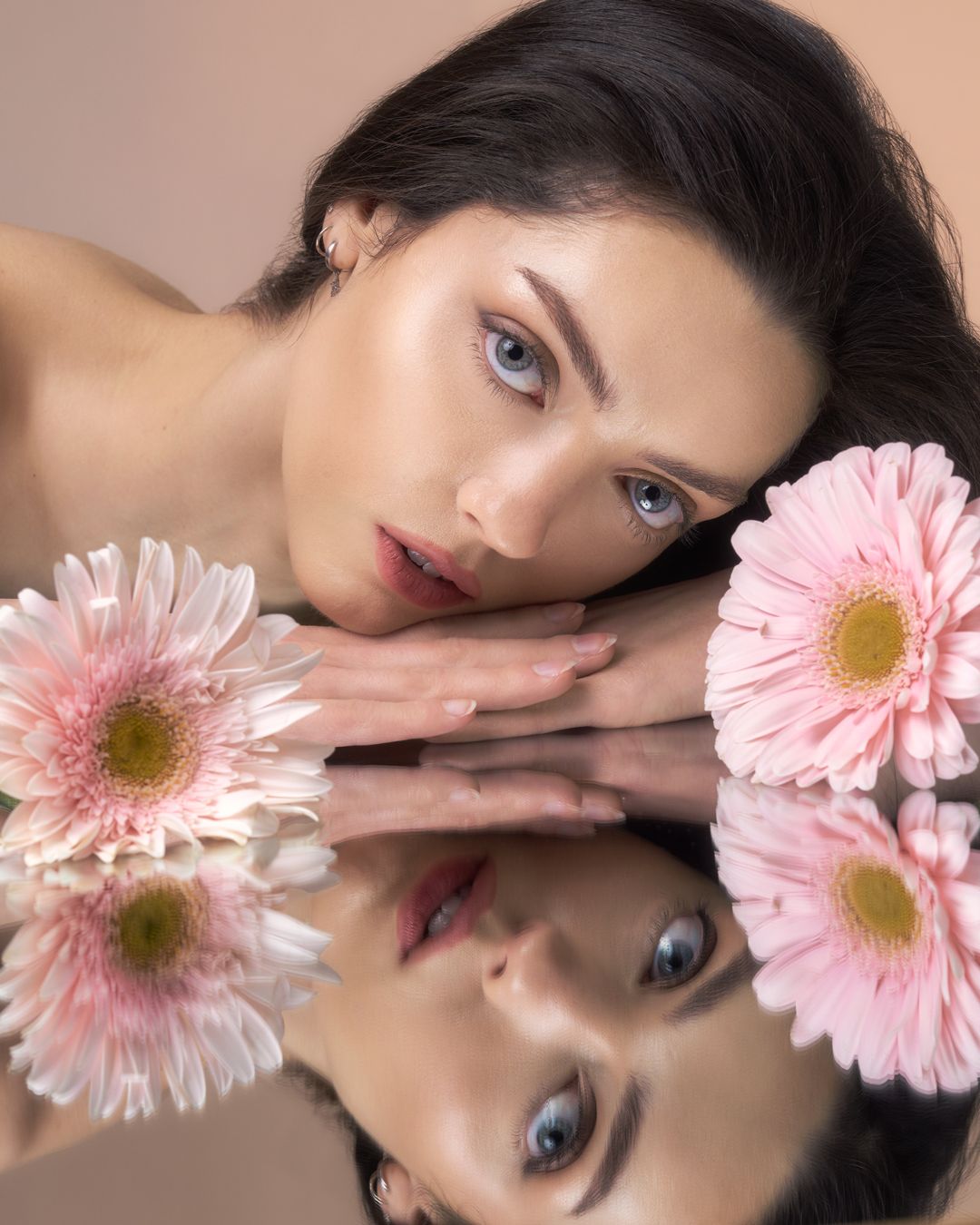 Young woman with blue eyes resting her face on hands beside pink gerbera daisies reflected on a mirror surface.