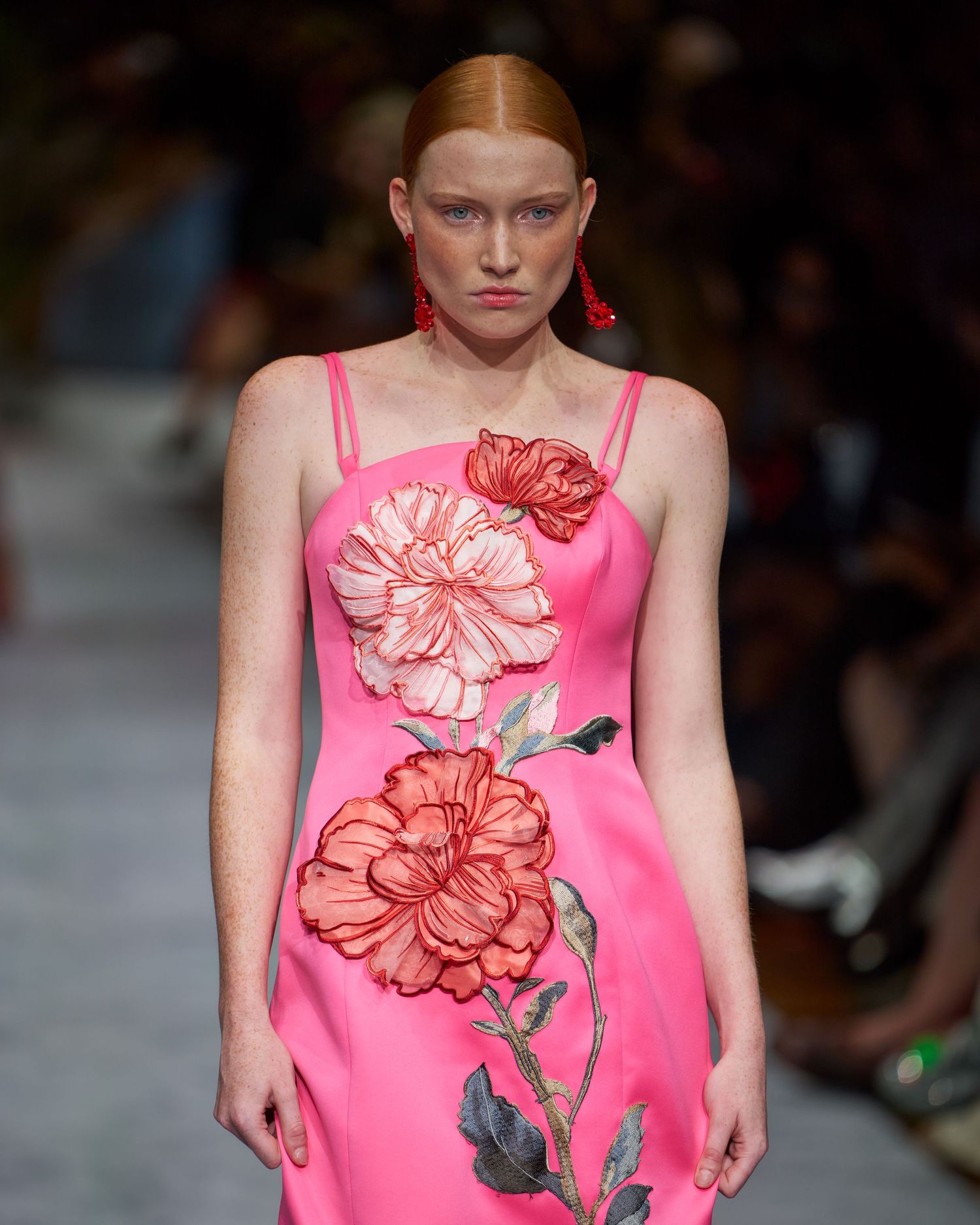 Pink evening gown with large floral appliques in coral and peach tones walks down a fashion runway.